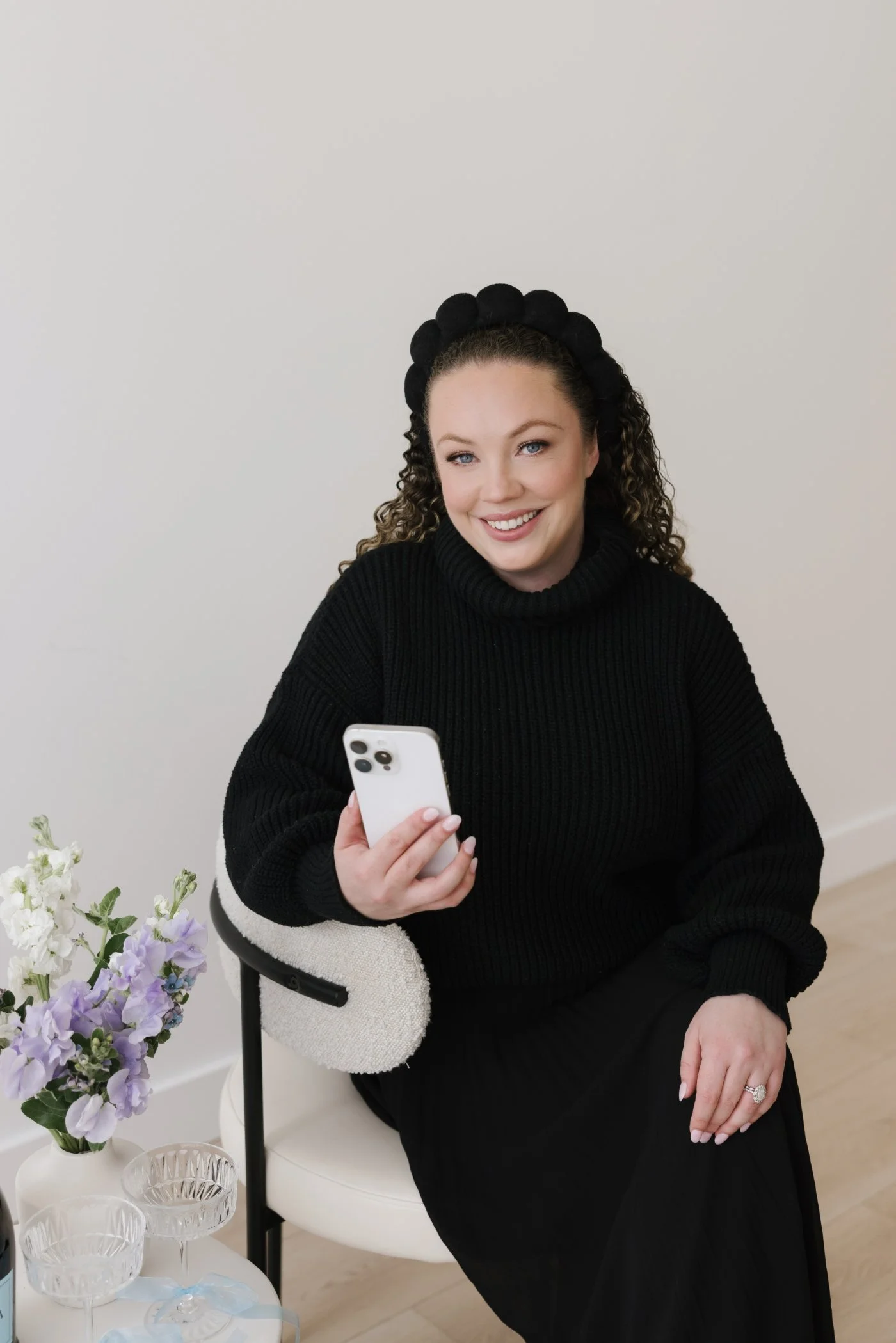 Cassie sitting a white chair, smiling and scrolling on her white iPhone.