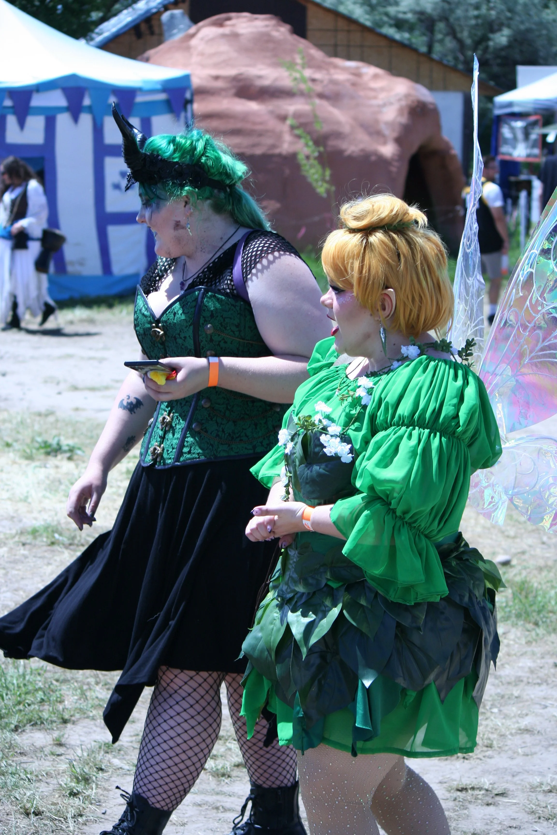 Two women dressed in colorful costumes and fairy wings talking and walking outdoors at a festival or fair, with tents and a rocky structure in the background.