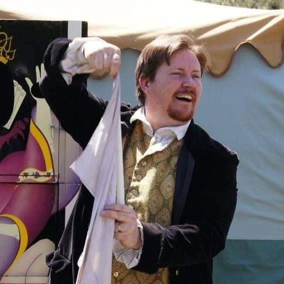 A man with light brown hair, wearing a patterned vest and white shirt, joyfully waving a white cloth outdoors.
