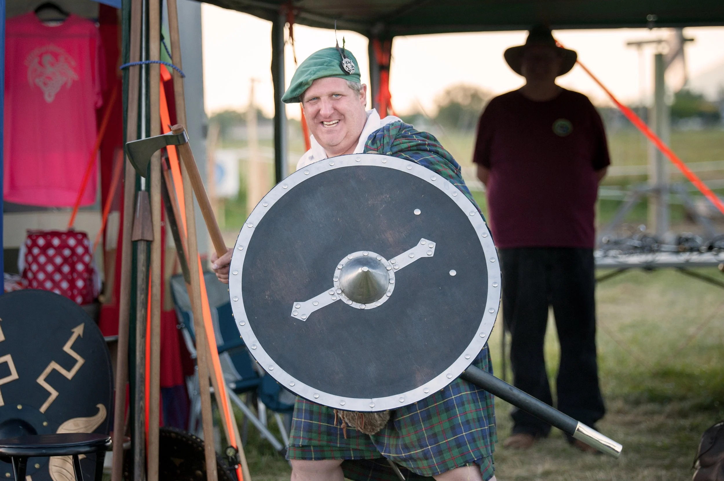 A man dressed in Scottish attire, including a green beret and plaid kilt, holding a round shield and axe, smiling at an outdoor event, with another person standing in the background under a tent.