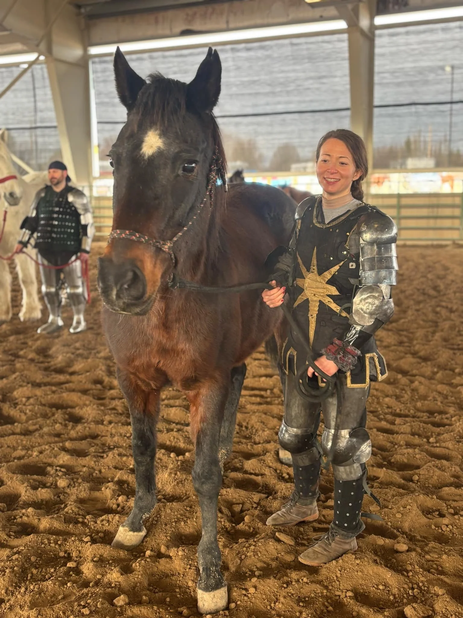 A woman dressed in medieval knight's costume standing next to a brown horse inside an indoor riding arena with another person in knight armor in the background.