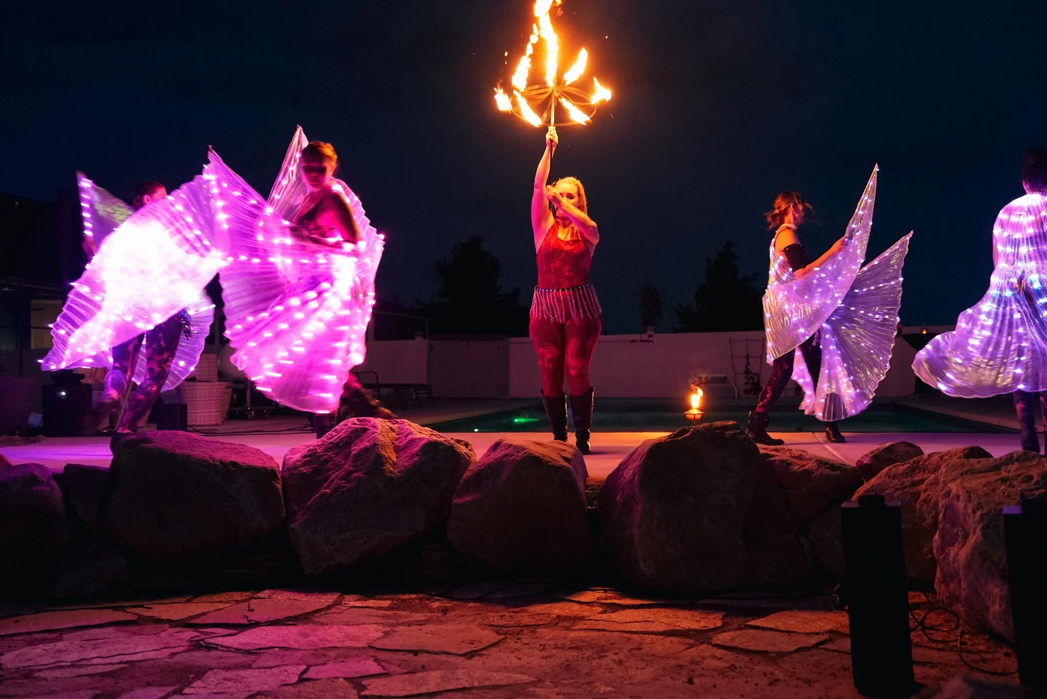 Performers dancing on stage at night with LED props; one woman in the center holds a flaming torch above her head, and others hold illuminated wings.