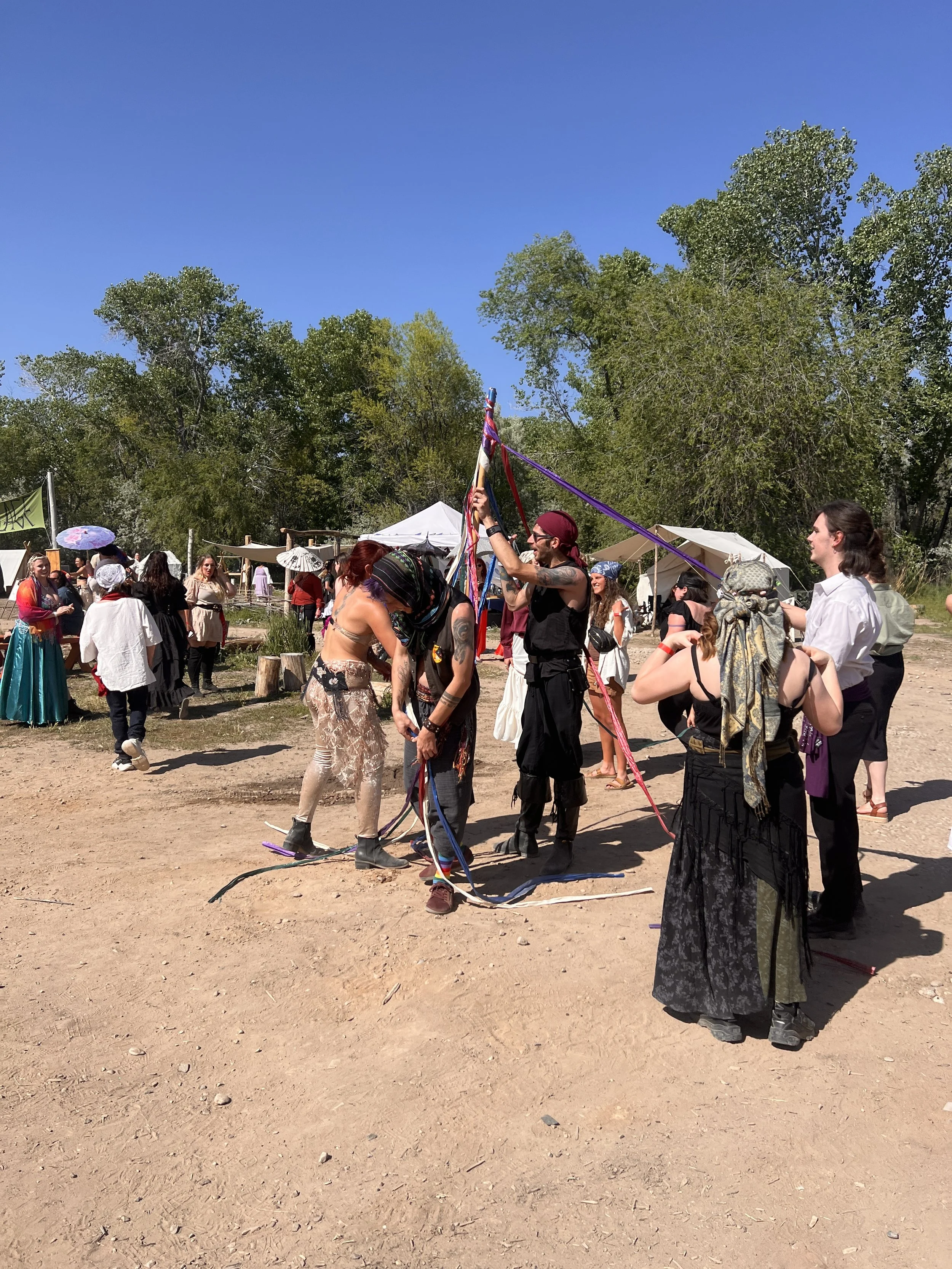 Group of people dressed in bohemian and festival attire dancing and socializing outdoors at a festival with trees and tents in the background.