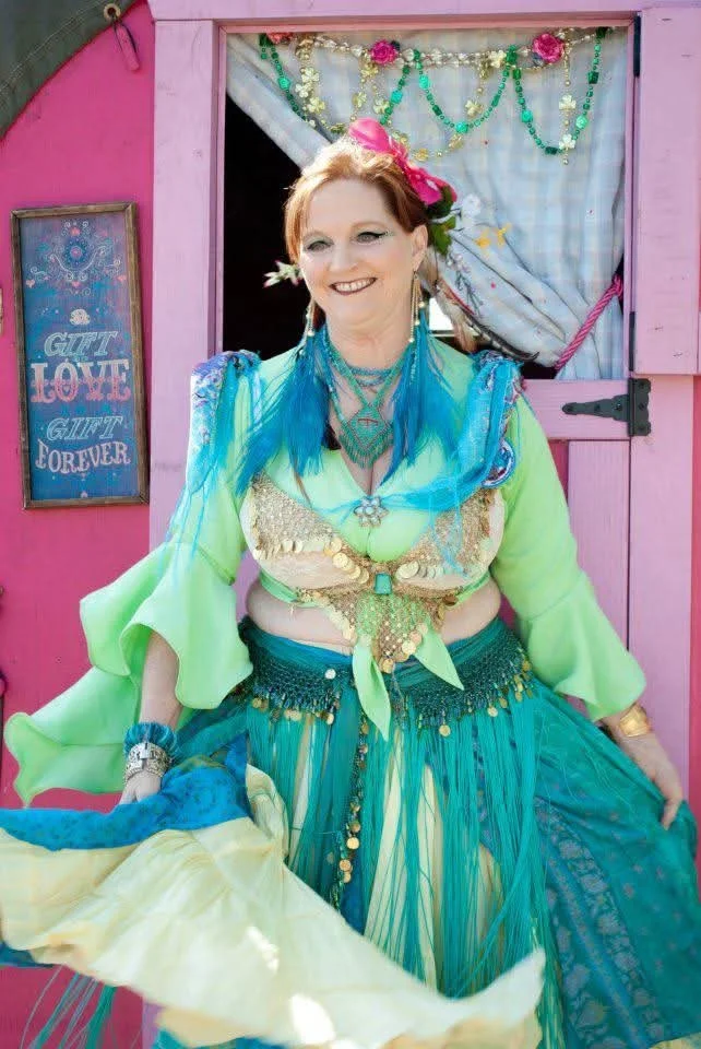 A woman dressed in colorful, layered belly dance costume with blue and green hues, bead and coin embellishments, standing in front of a pink door decorated with beads and flowers, smiling at the camera.