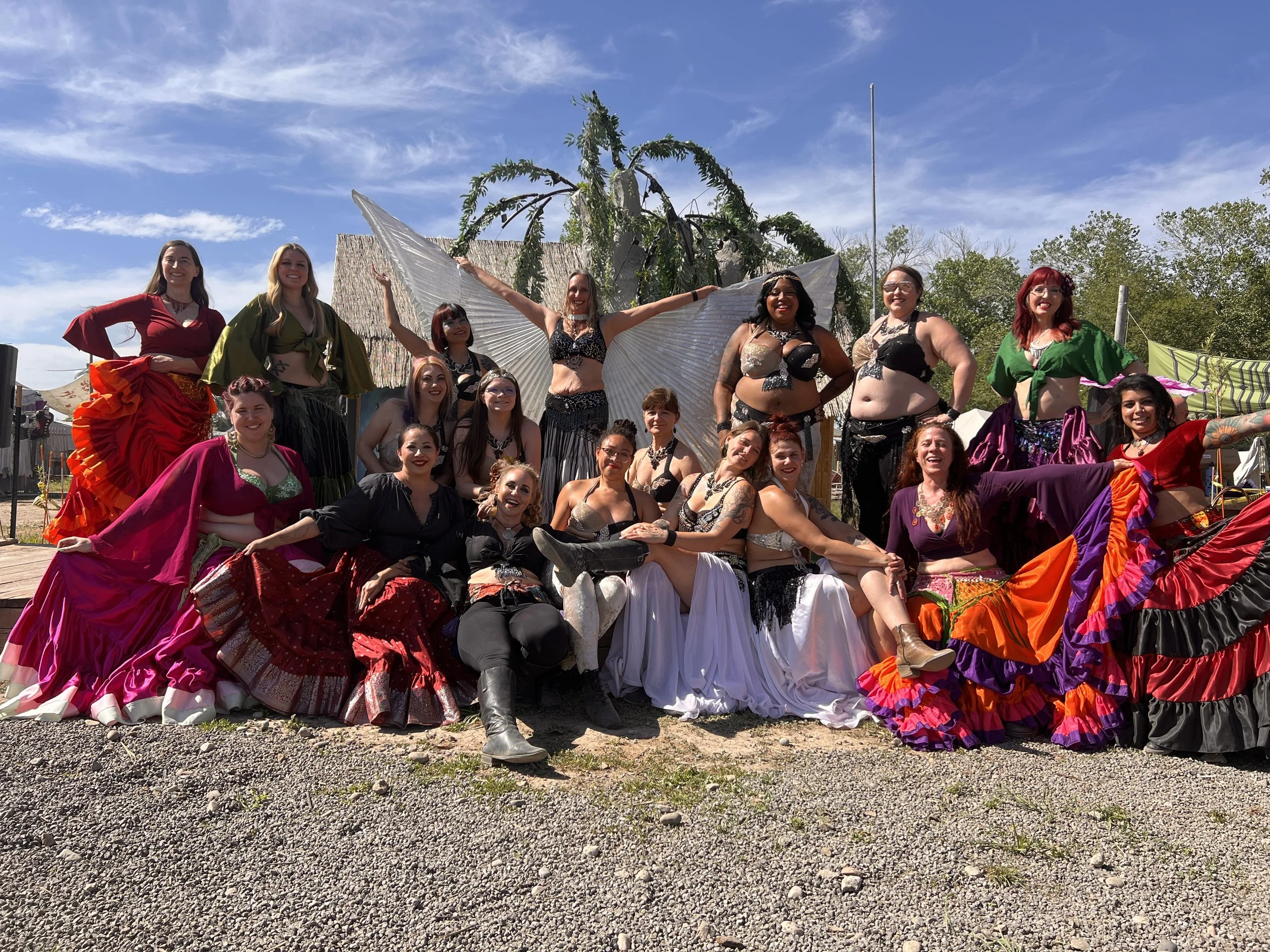 Group of women in colorful vintage-style dresses posing outdoors on a sunny day with trees and tents in the background.