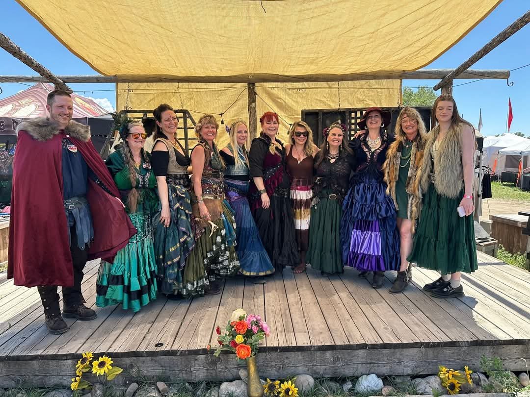 A group of people dressed in medieval or fantasy costumes posing on a wooden stage under a yellow canopy at an outdoor event.