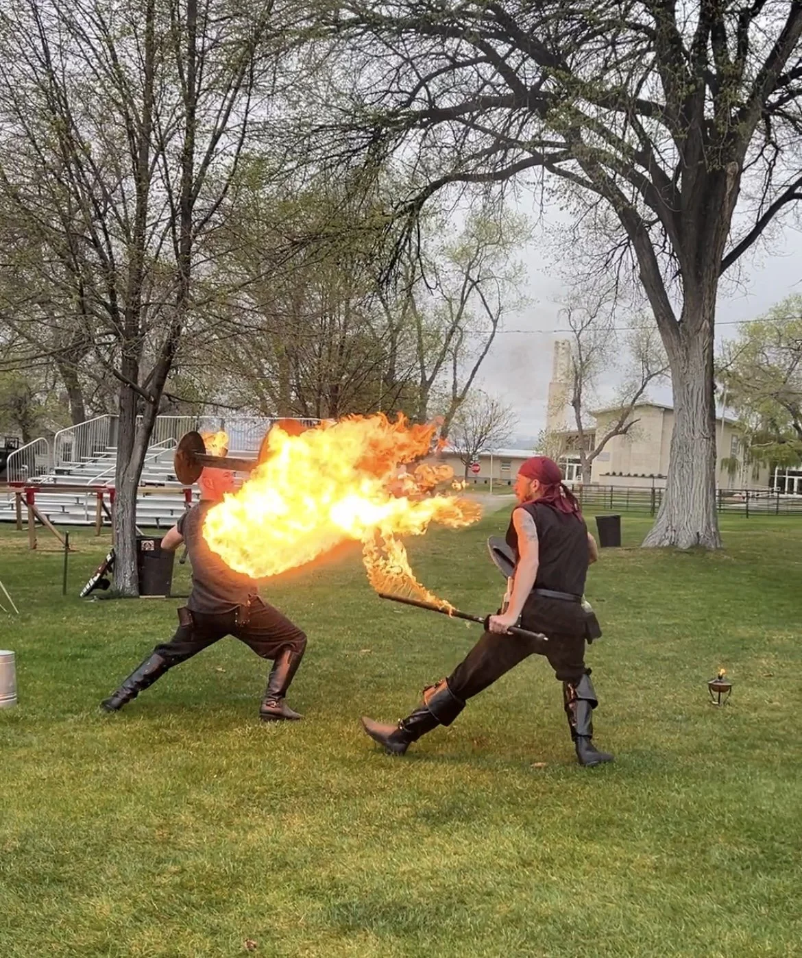Two men performing a fire-breathing and fire-fighting stunt outdoors with flames, trees, and a cloudy sky in the background.