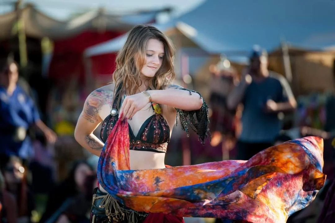 Young woman with tattoos, wearing a black top with colorful patterns, weaving vibrant fabric at outdoor market.