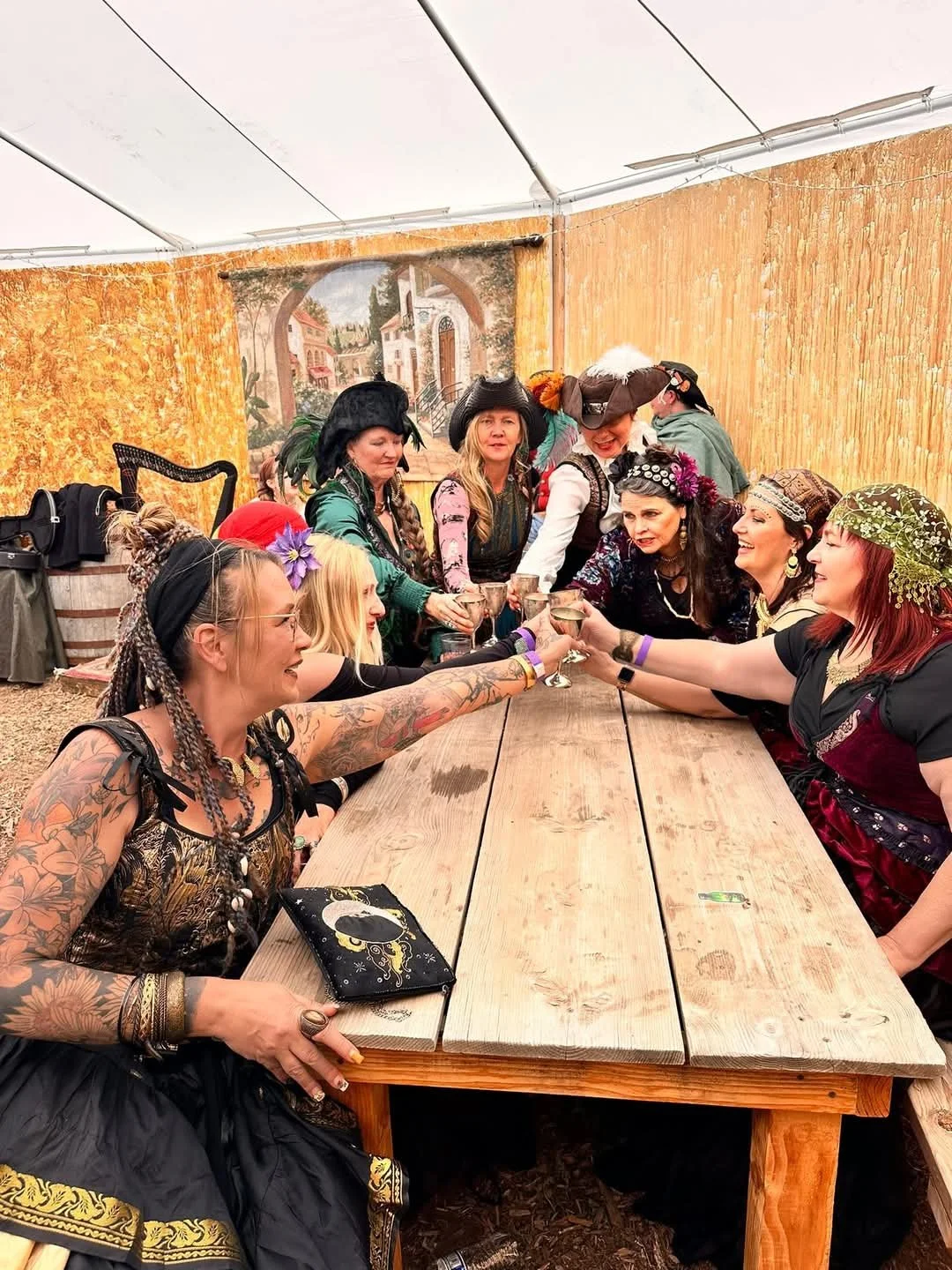 Women dressed in pirate costumes sitting around a wooden table, raising glasses in a toast at a themed party under a tent.