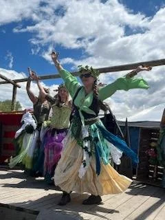 Group of women dressed in colorful traditional skirts and tops, performing a dance outdoors on a wooden platform under a partly cloudy sky.