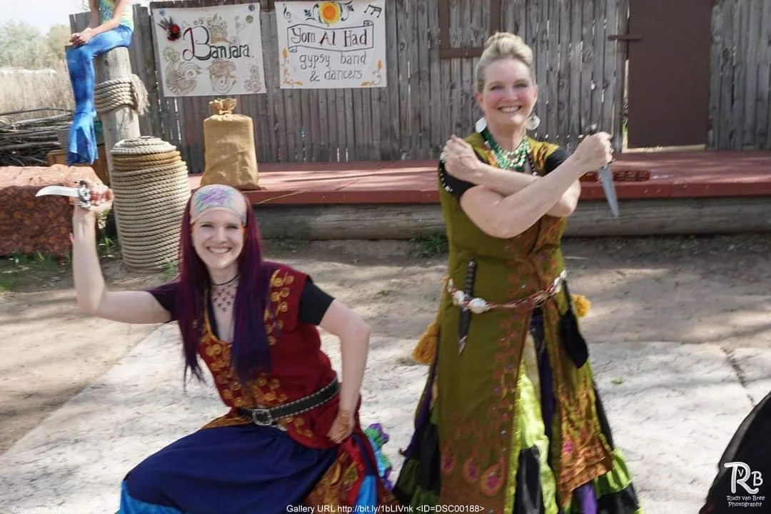 Two women dressed in pirate or gypsy costumes pose playfully outdoors, one holding a toy knife and the other crossing her arms with a toy sword. Behind them, a wooden fence has signs that read 'Banzaa' and 'You Al Had gypsy band & dancers'.