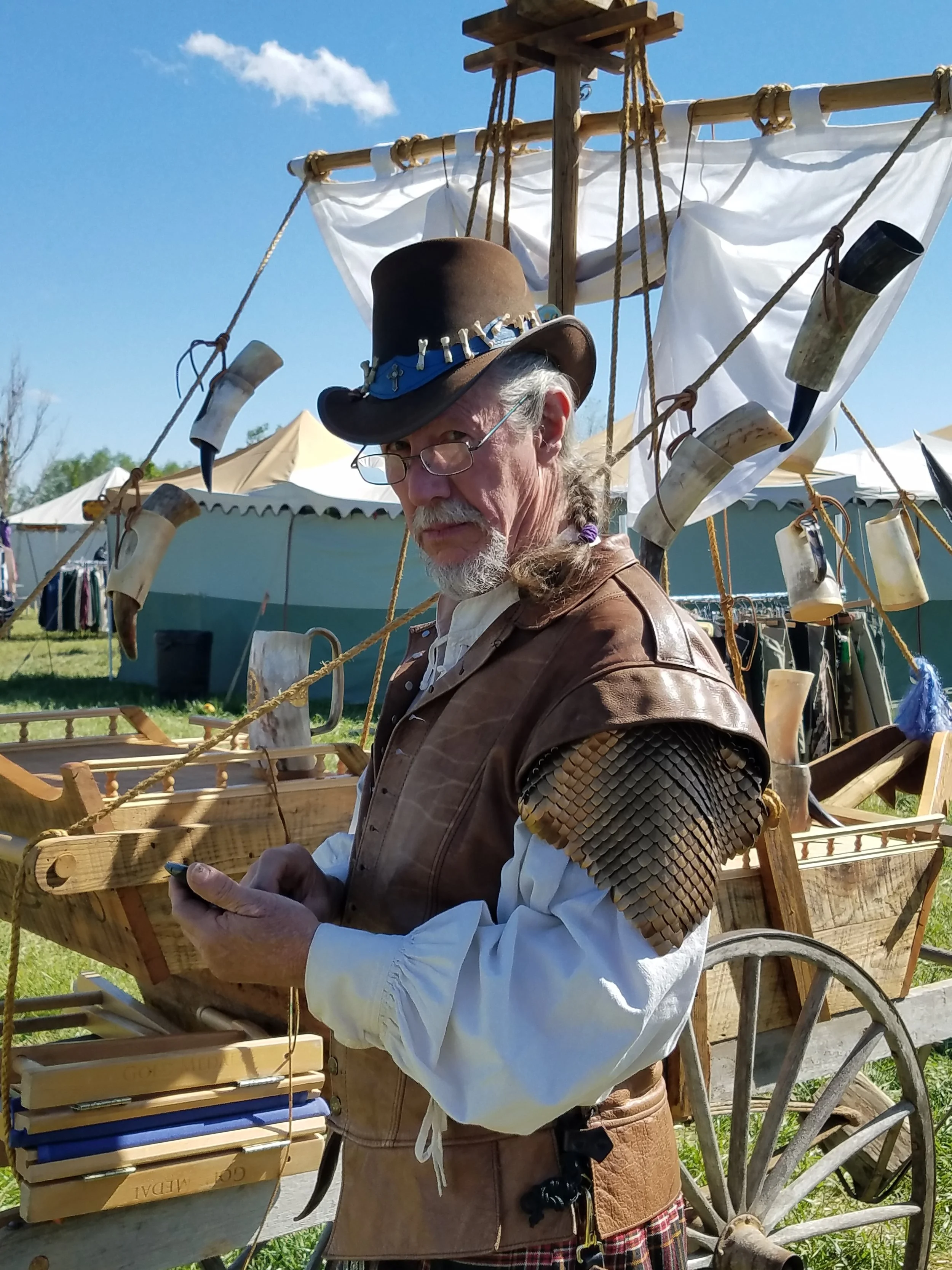 A man dressed as a pirate with a leather vest, fish scale armor on his shoulders, white shirt, and a wide-brimmed leather hat with metal embellishments. He is standing in front of a wooden cart with a pirate-themed ship decoration made of wood and fa