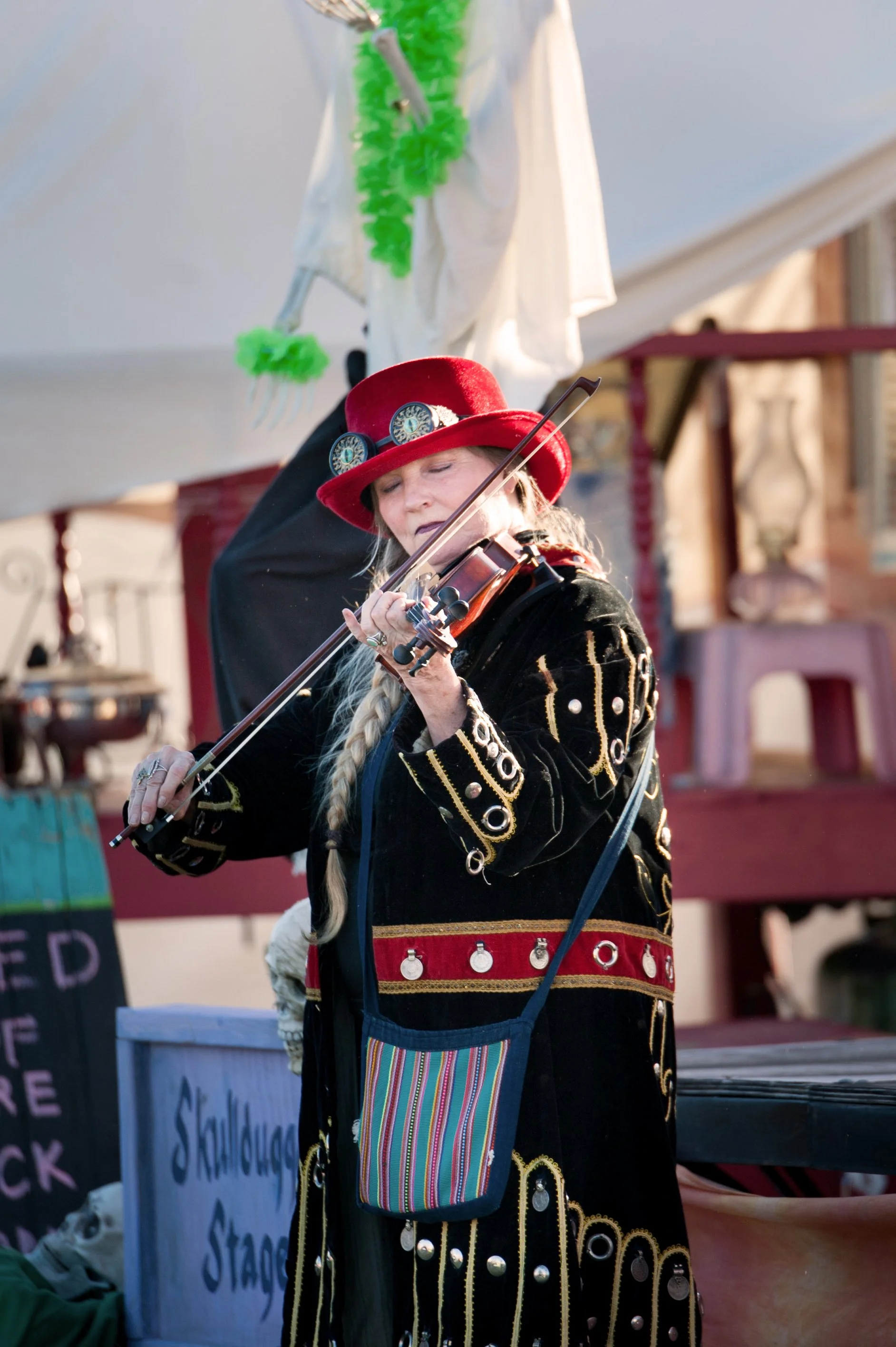 A woman playing the violin outdoors, wearing a black velvet jacket with gold and silver embellishments, a red wide-brimmed hat with decorative circles, and a colorful striped pouch hanging from her shoulder.
