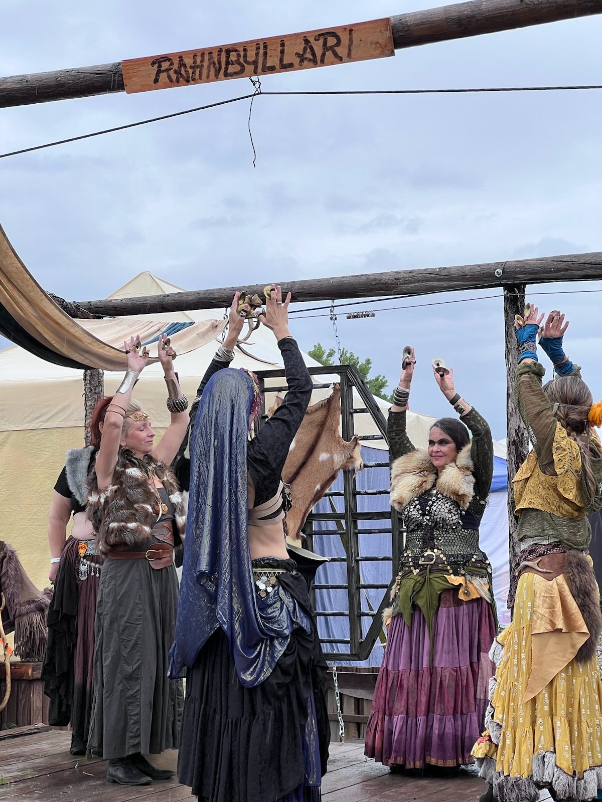 A group of people dressed in medieval or fantasy costumes, standing on a wooden stage with arms raised, at a renaissance fair or themed event. There is a large sign reading 'RAHNBULLARI' hanging above them.