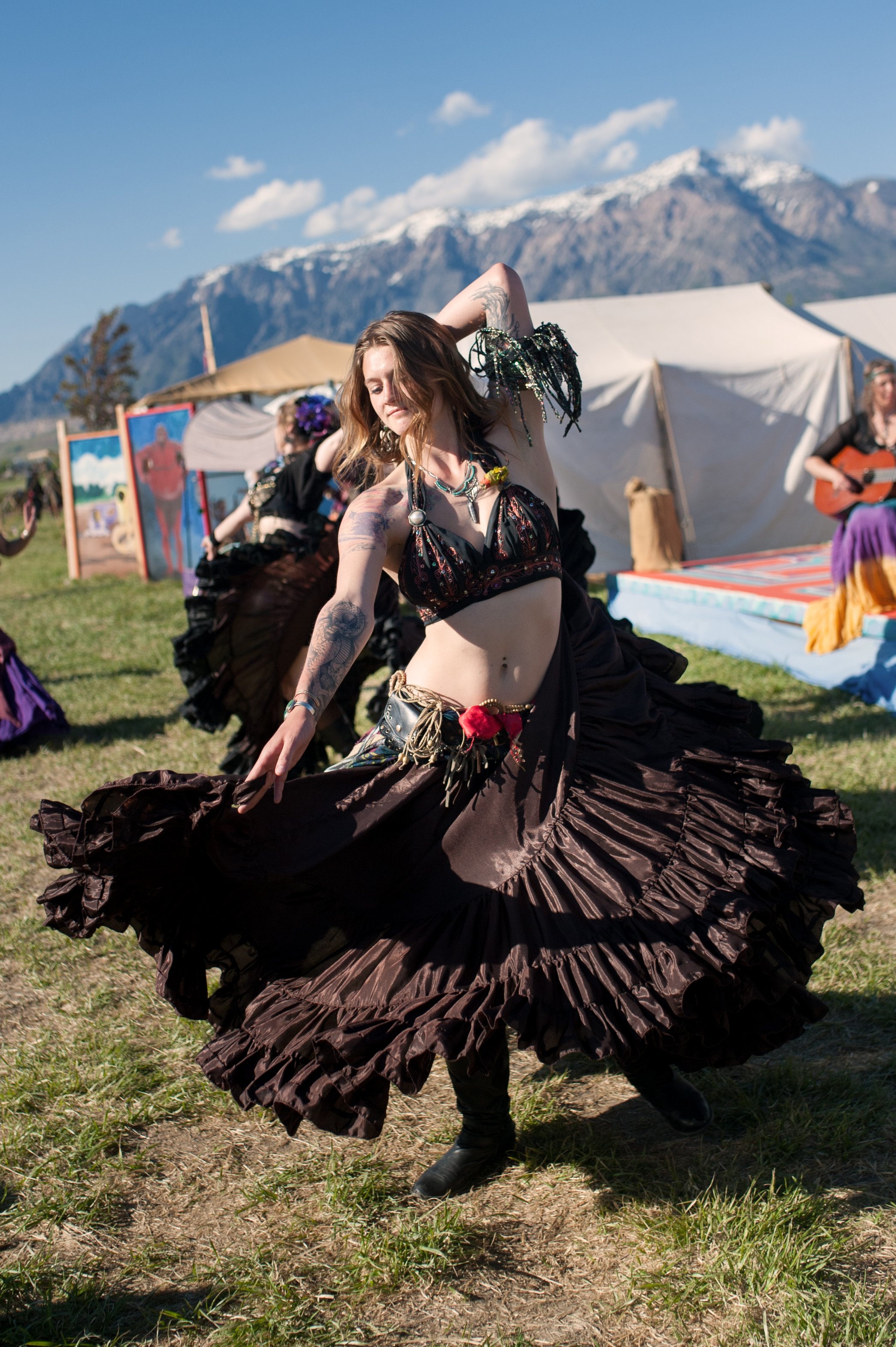 A woman with tattoos dancing in a brown skirt and black top at an outdoor festival with tents and mountains in the background.