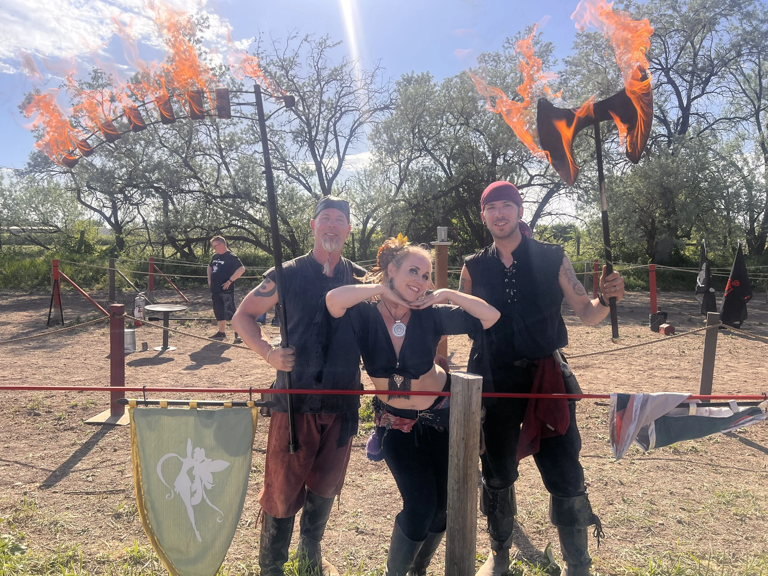 Three people dressed as medieval or fantasy characters, one woman and two men, at an outdoor event with flames and banners in the background. They are smiling and posing for the photo.