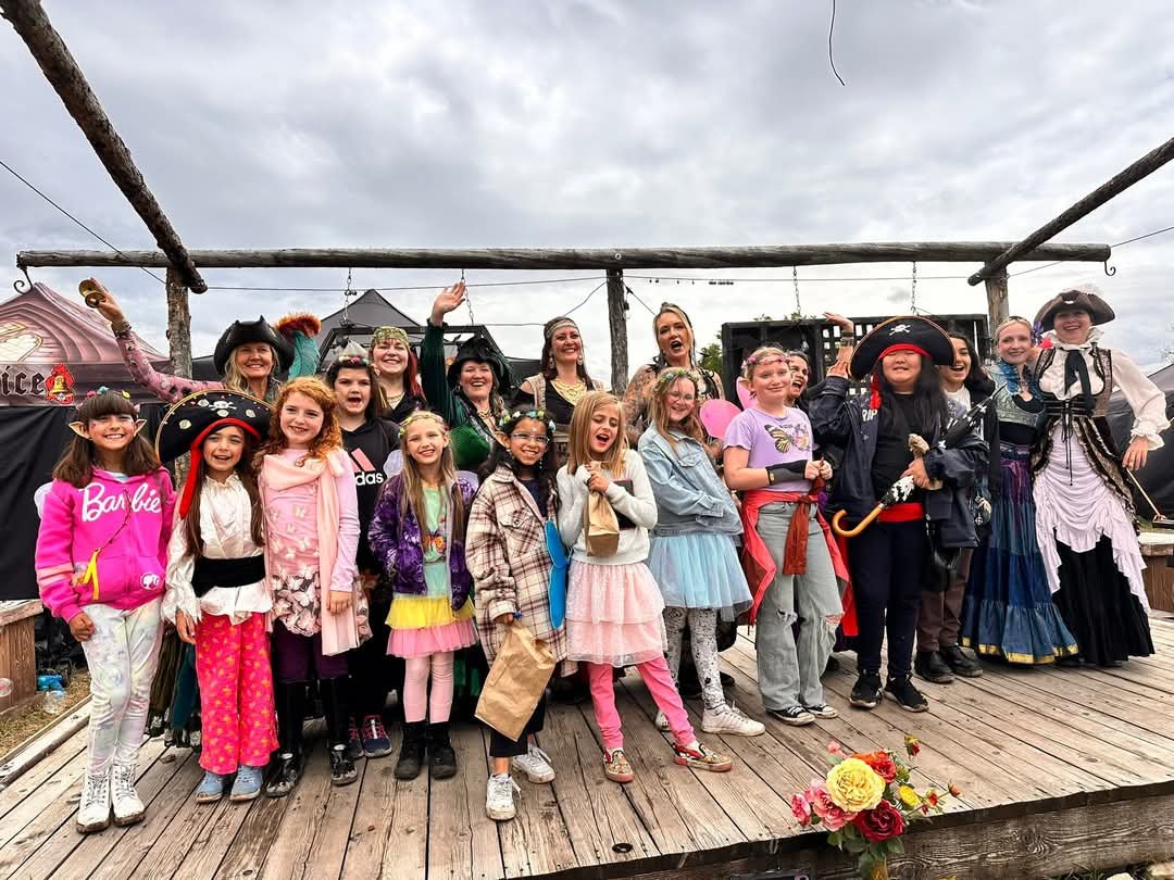 Group of children and women dressed as pirates and princesses on a wooden stage outdoors under cloudy sky.