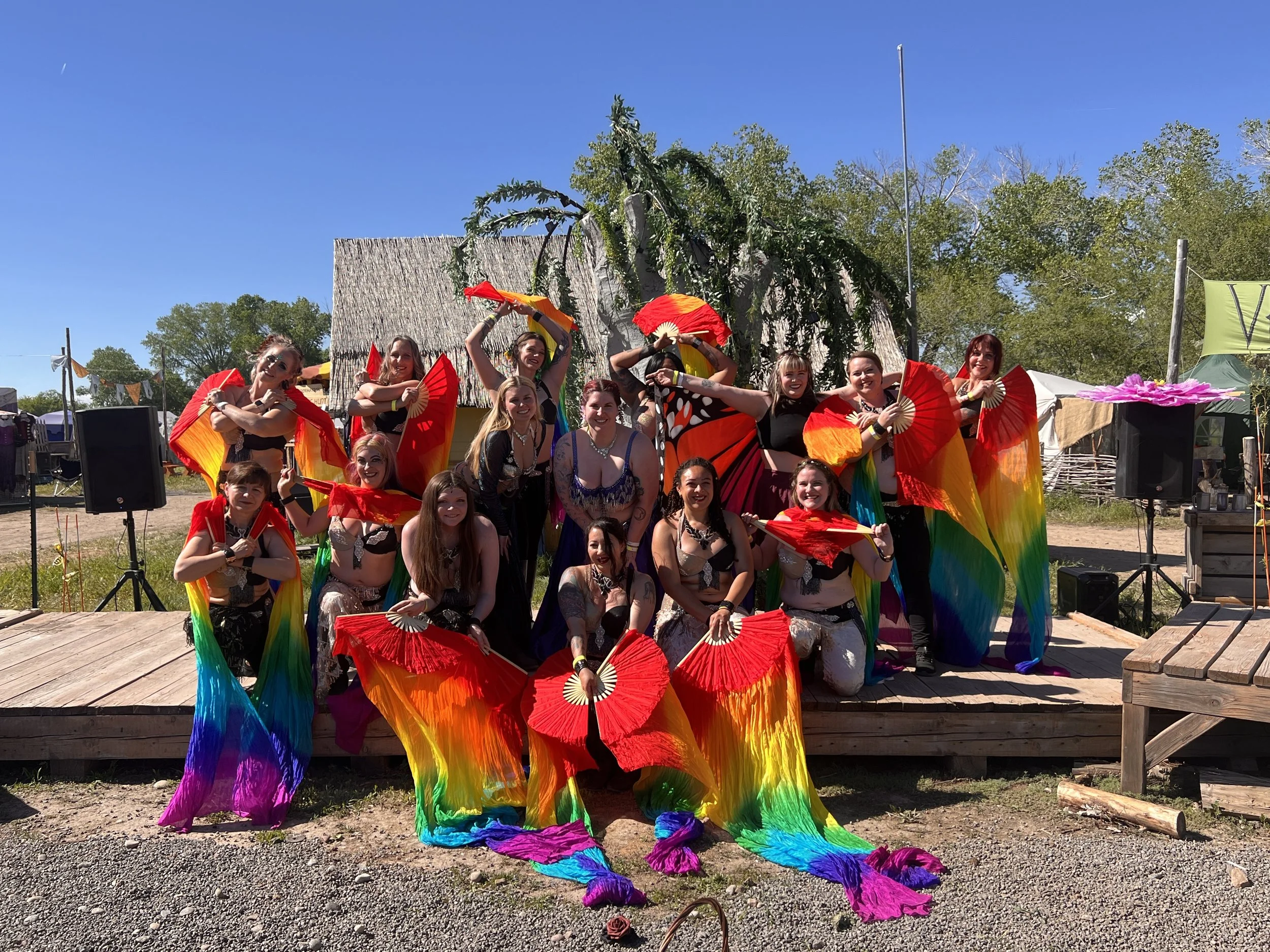 Group of dancers in colorful costumes holding fans, posing outdoors on a wooden stage with a thatched roof structure in the background, under a bright blue sky with trees around.