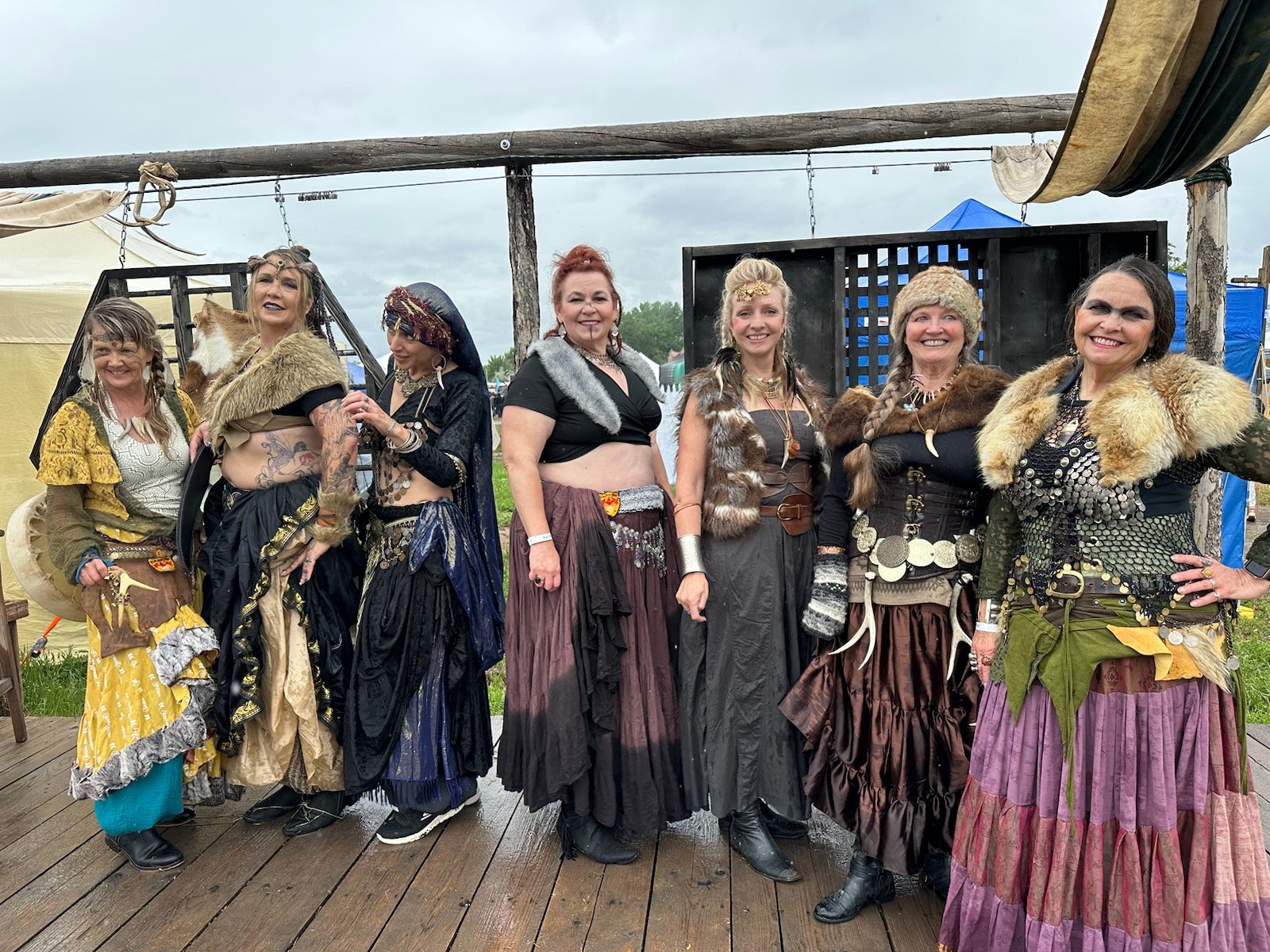Group of women dressed in fantasy or medieval costumes with various fur, leather, and fabric accessories standing on a wooden platform outdoors.