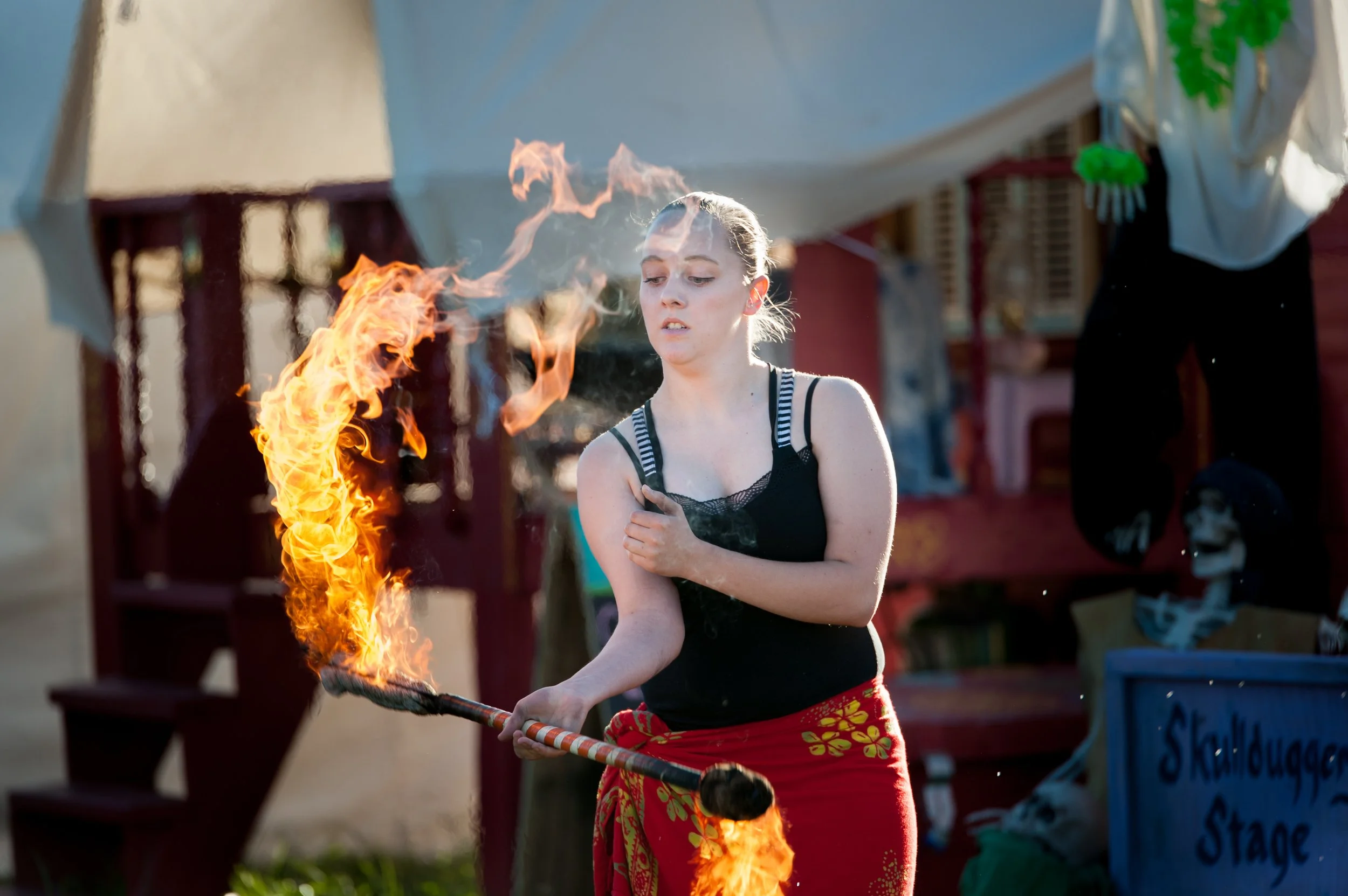 A young woman performing a fire dance, holding a flaming stick, with a focused expression. She wears a black top and a red skirt with yellow floral patterns, with an outdoor setting and other carnival or circus props in the background.