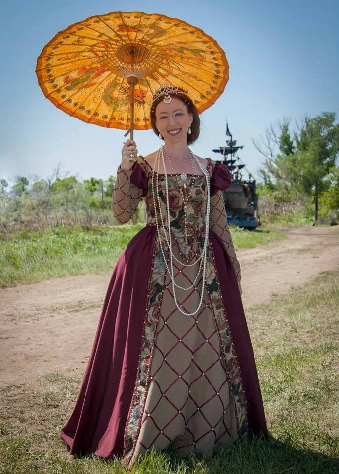 Woman dressed in a historical gown, holding a decorative parasol, standing outdoors on a dirt path with green trees and a building with a pagoda-style roof in the background.