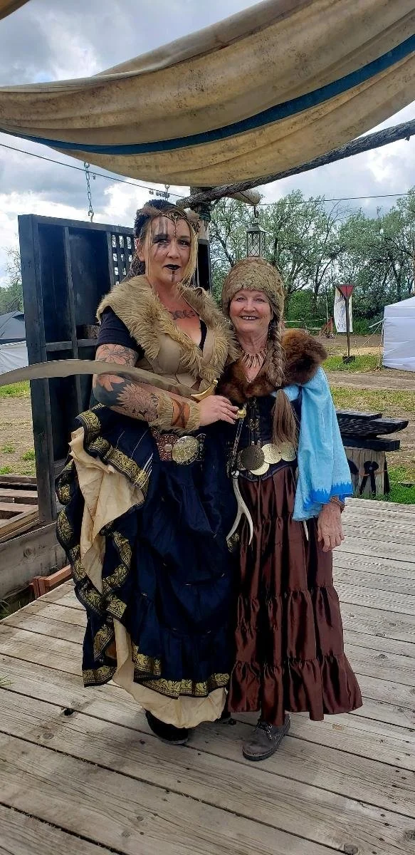 Two women in pirate costumes standing outdoors on a wooden platform, smiling at the camera. One woman has long hair, dark makeup, and a black pirate dress with a fur collar. The other woman has a brown braid, a tan knit hat, and a brown skirt with a 