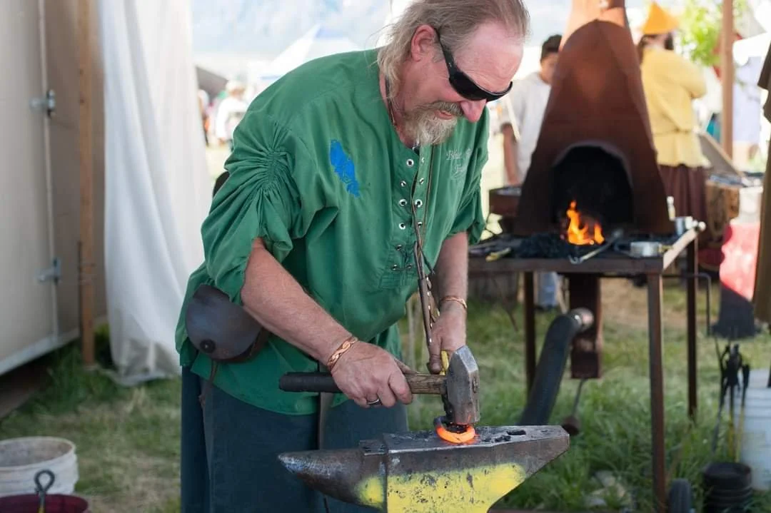A man wearing sunglasses and a green shirt smithing a piece of orange-hot metal on an anvil outdoors at a craft fair or festival, with a forge burning in the background.
