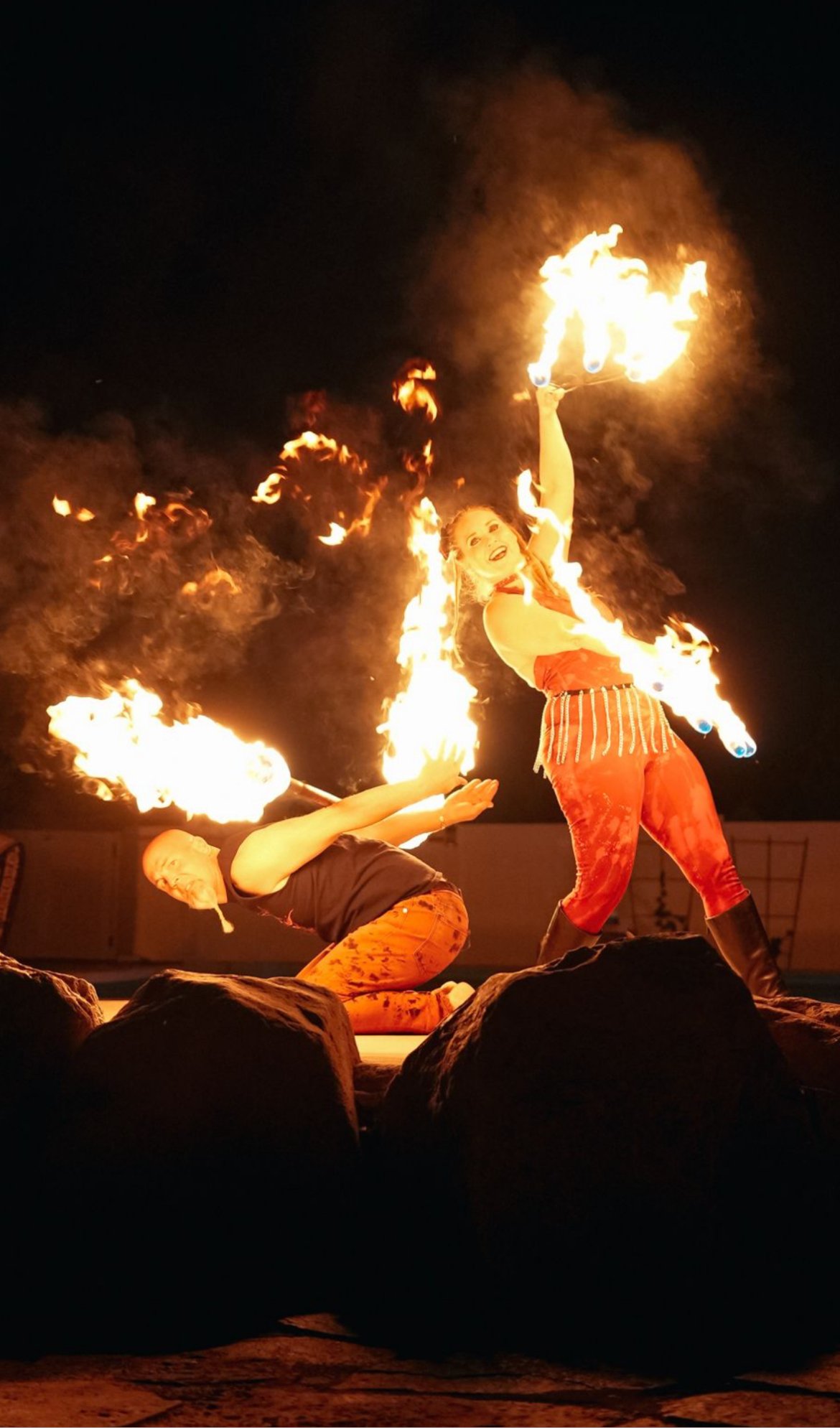 Two fire performers entertaining outdoors at night, one woman spinning fire and one man lying on the ground with fire in his mouth.