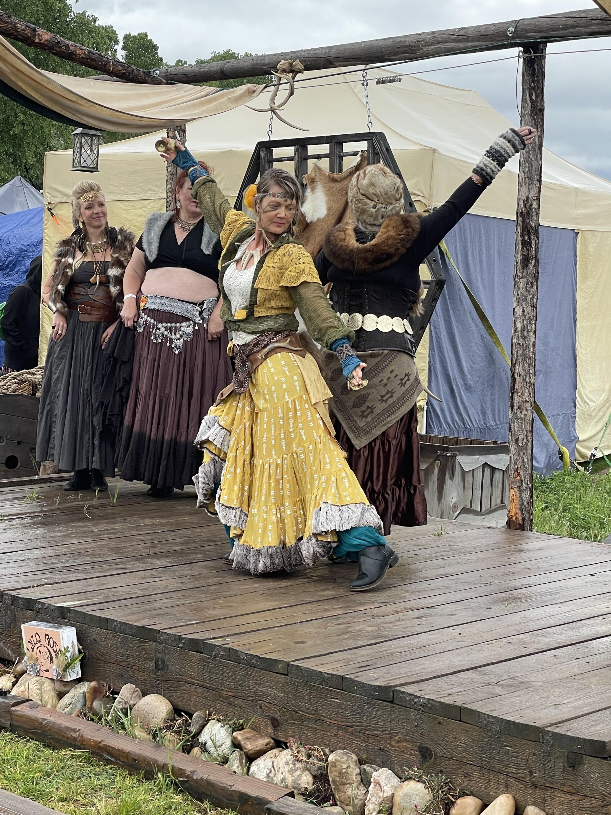 A group of people dressed in medieval or fantasy costumes dancing on a wooden stage outdoors at a festival or reenactment event.