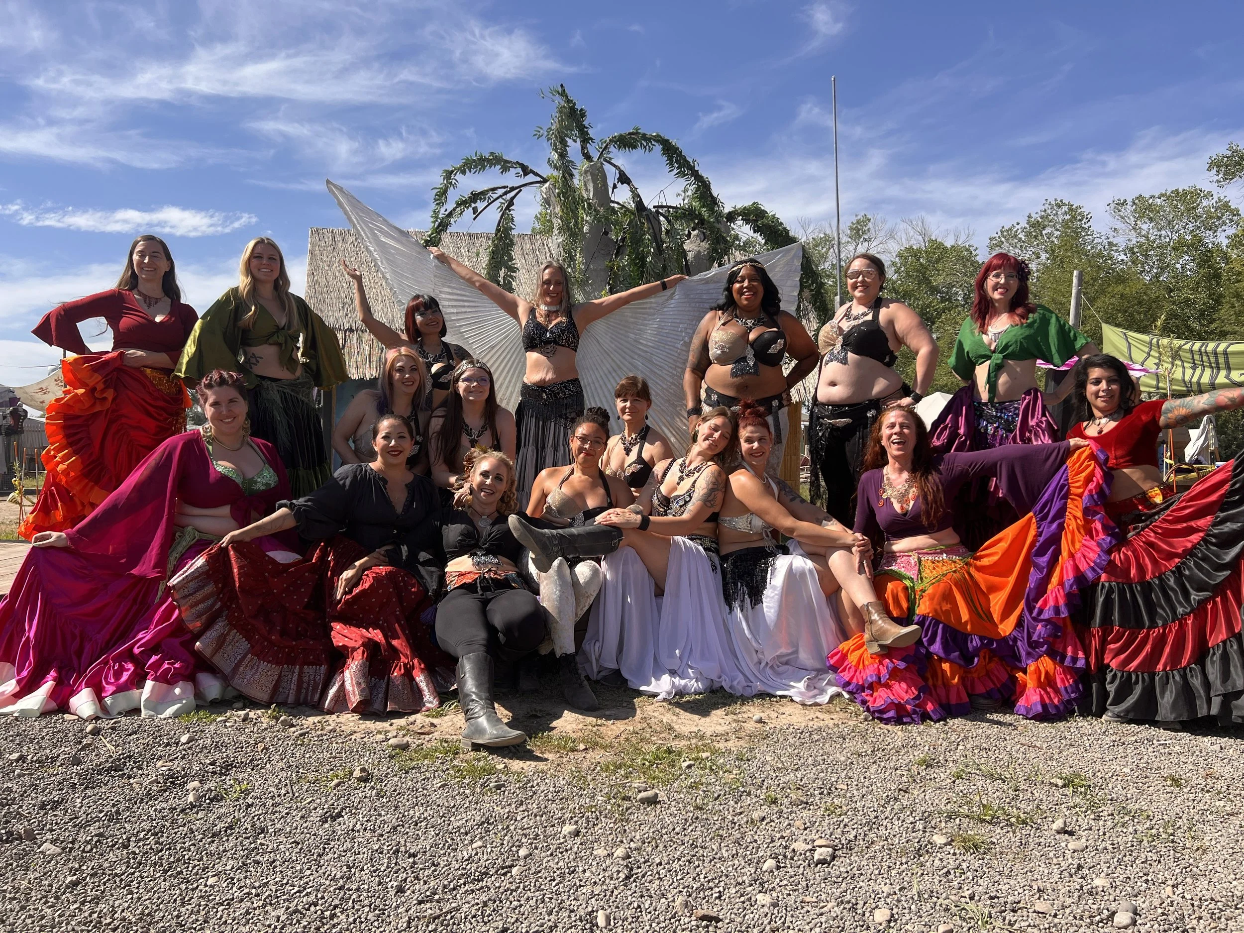 Group of women in colorful belly dance costumes posing outdoors on a sunny day, with a large decorative backdrop and trees in the background.