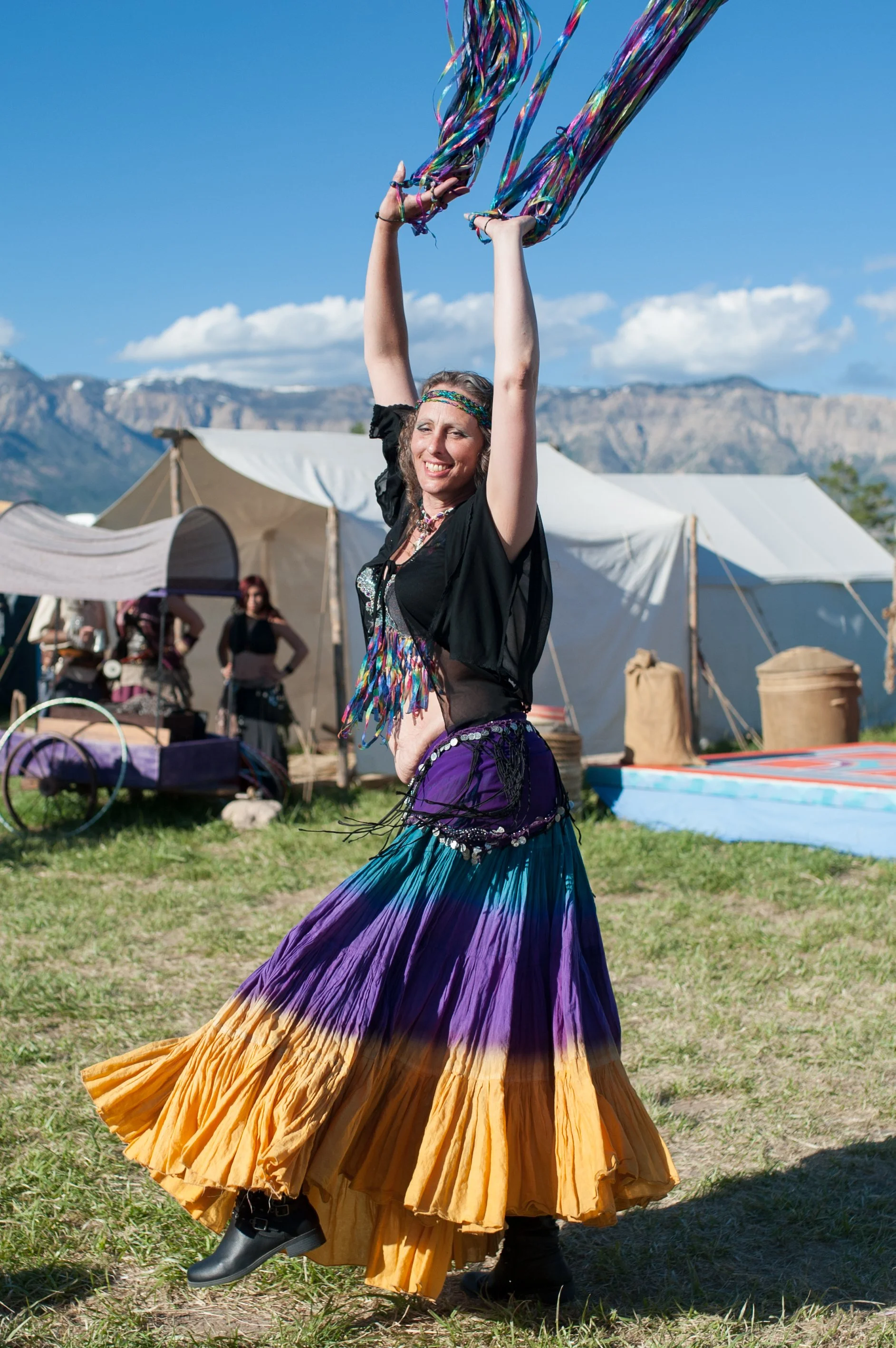 A woman wearing a colorful, flowing skirt and black top dancing outdoors during a festival, holding streamers high above her head with mountains and festival tents in the background.