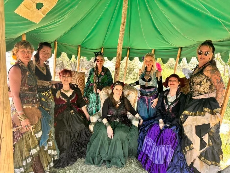 Group of women in elaborate, colorful pirate-themed or fantasy costumes, seated and standing inside a green tent outdoors.