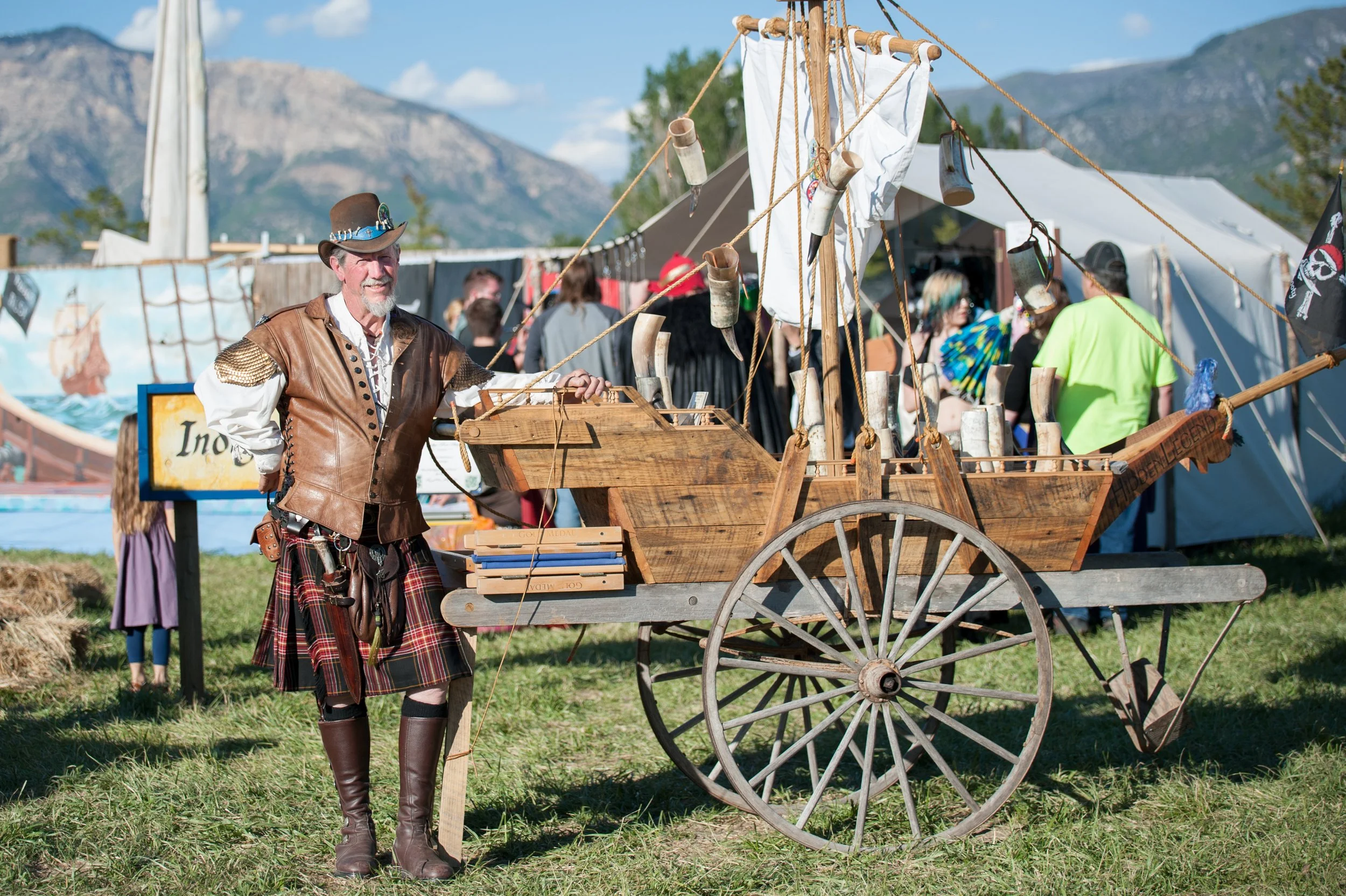 A man dressed as a pirate in a leather vest, plaid kilt, and tall leather boots stands next to a wooden ship replica on a cart at a fair or festival, with tents, people, and mountains in the background.