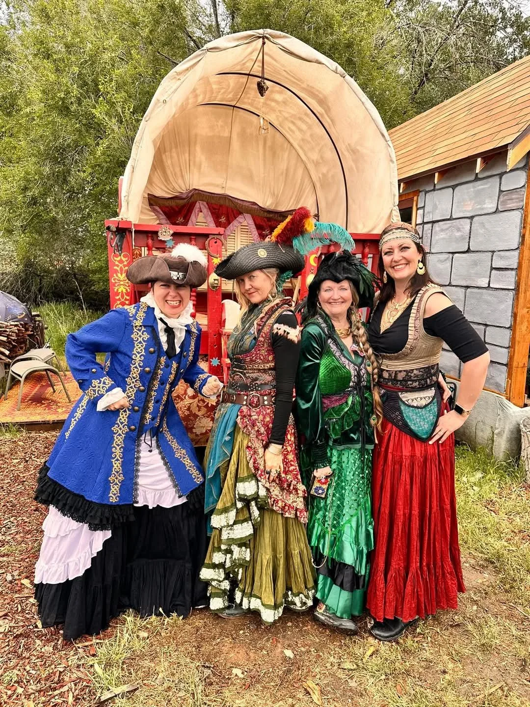 Four women dressed in colorful pirate costumes, standing in front of a red carriage with a beige canopy, outdoors near a gray stone building.