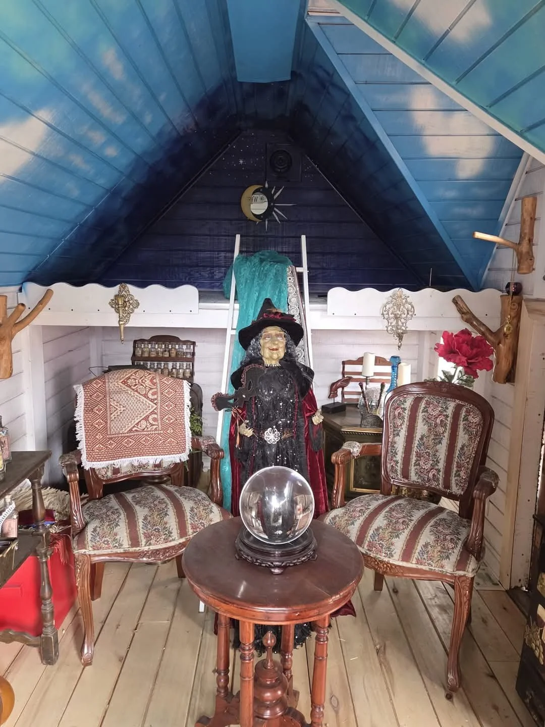 A cozy attic room decorated with vintage furniture, including two patterned armchairs, a small round wooden table with a glass globe, a witch figurine, a ladder with a teal cloth, and various decorative items on the walls and shelves. The ceiling is 