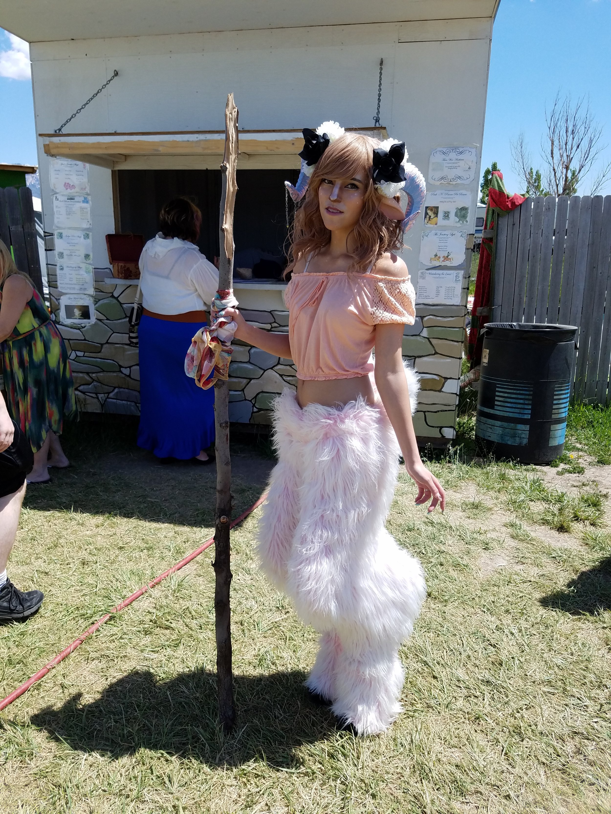 A young woman dressed as a fantasy creature with horns and furry pants, standing outdoors on grass, holding a staff, at a fair or festival.