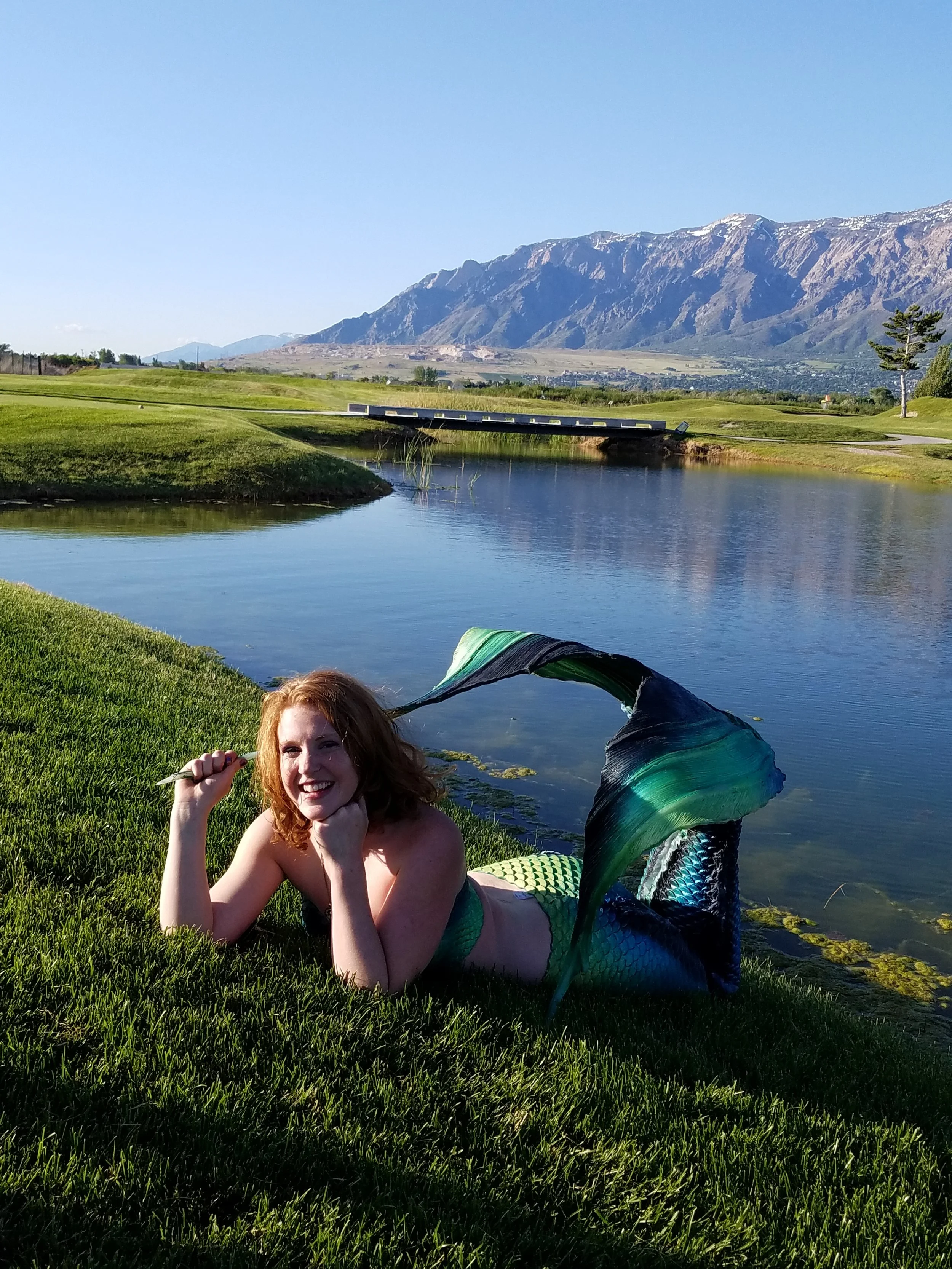 A woman dressed as a mermaid lying on the grass by a lake, smiling and holding a stick; mountains and a bridge are visible in the background on a clear day.