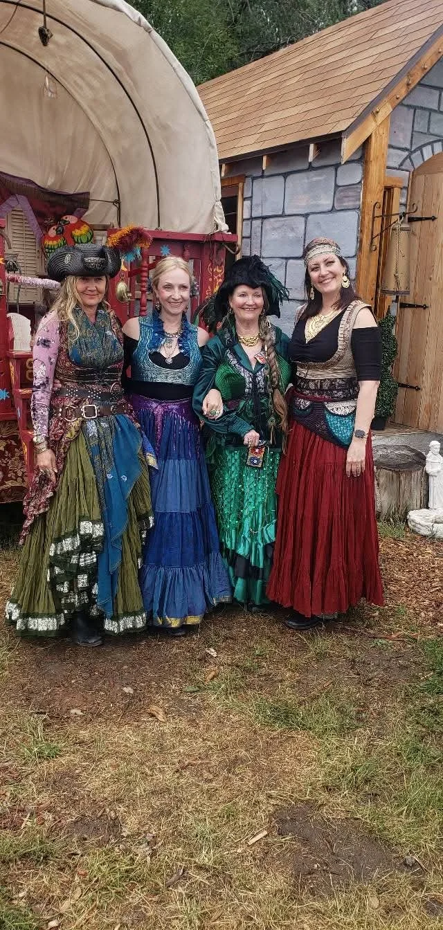 Four women dressed in colorful, elaborate costumes posing outdoors in front of a small wooden and stone building.