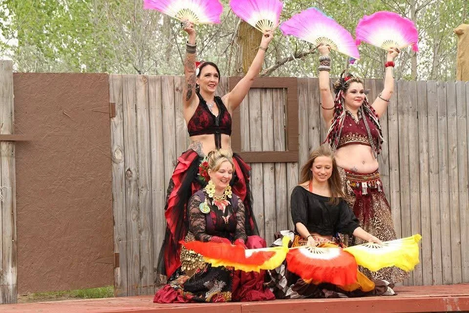 Four women in colorful, traditional belly dance costumes performing outdoors in front of a wooden fence. Two women at the front are sitting on the ground with large, vibrant fans, while two women at the back are standing, holding similar fans up in t