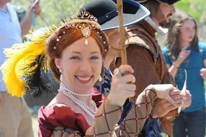 A woman dressed in historical costume with a feathered hat, holding a sword and smiling at historical reenactment event.