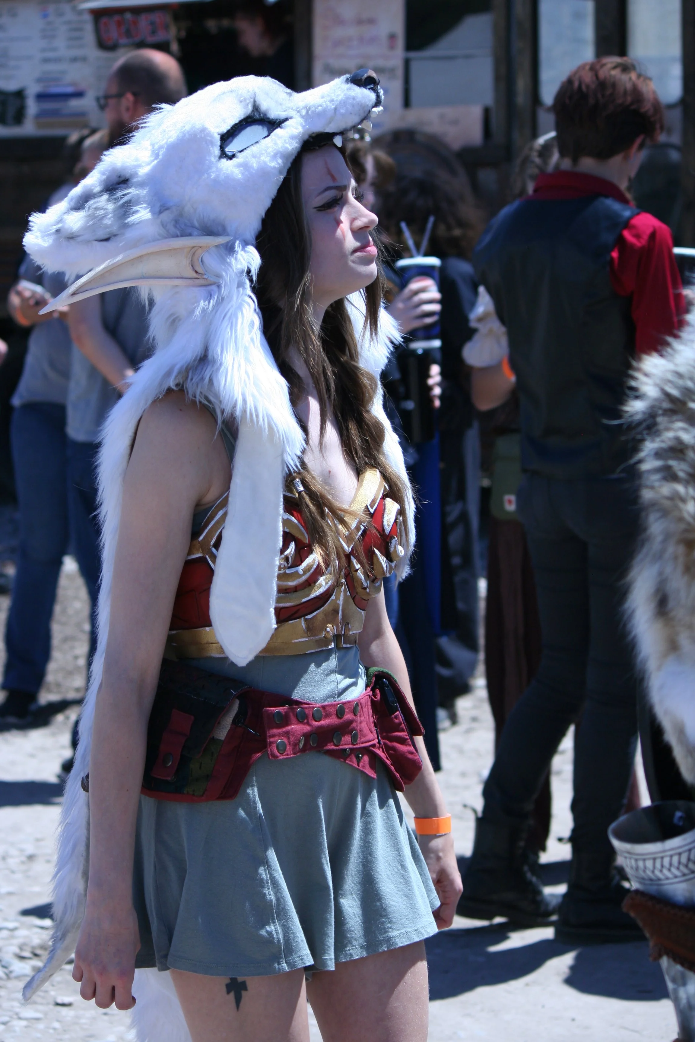 Person wearing a wolf head costume, a red corset, and a gray skirt, standing among a crowd outdoors.