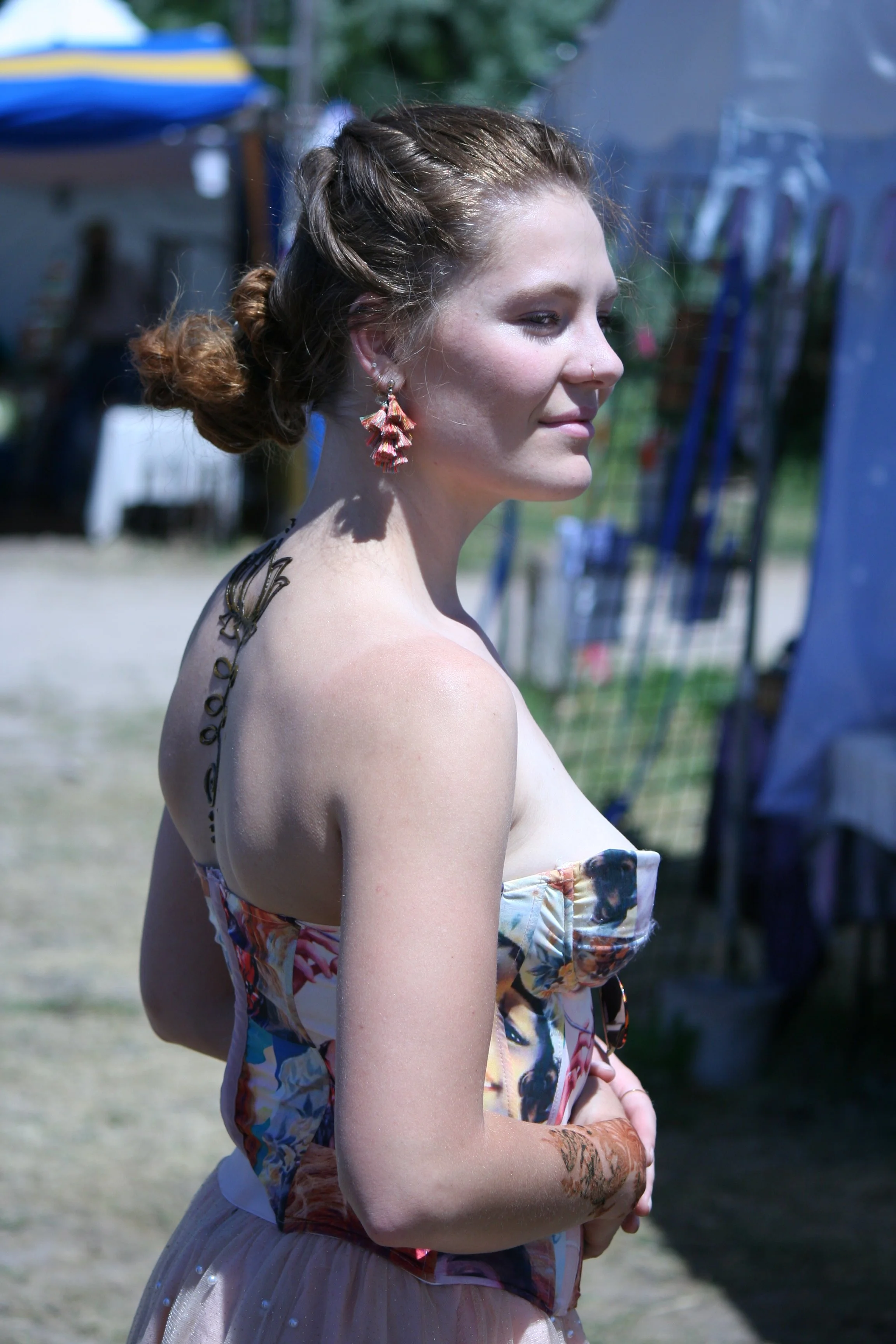 A woman with red hair styled in an updo, wearing dangling pink earrings, a strapless floral dress, and visible tattoos on her shoulder and arm, standing outdoors on a sunny day with a blurred background of tents and greenery.