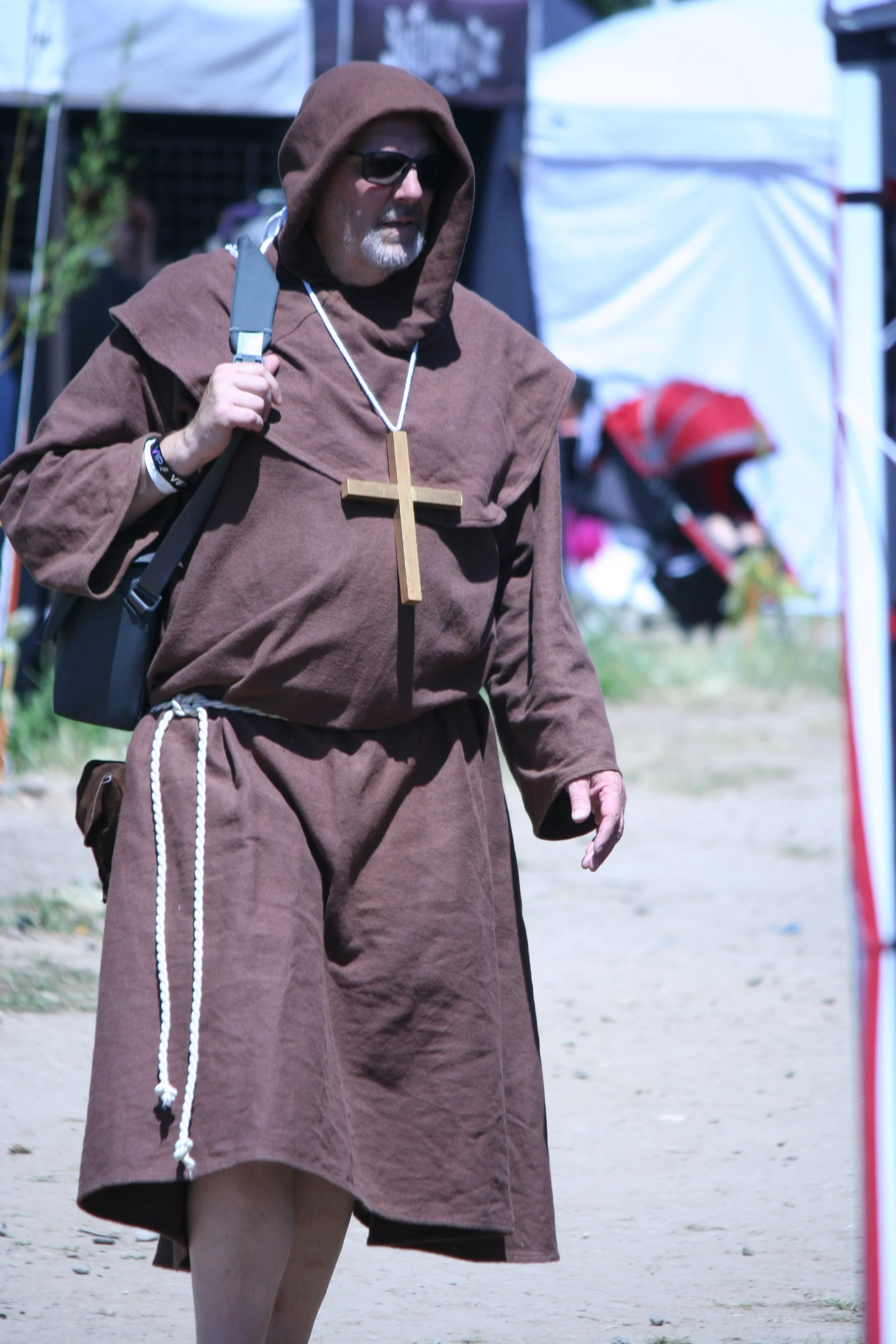 A person dressed as a monk with a brown robe, hood, and a large wooden cross necklace, holding a backpack strap, walking outdoors.
