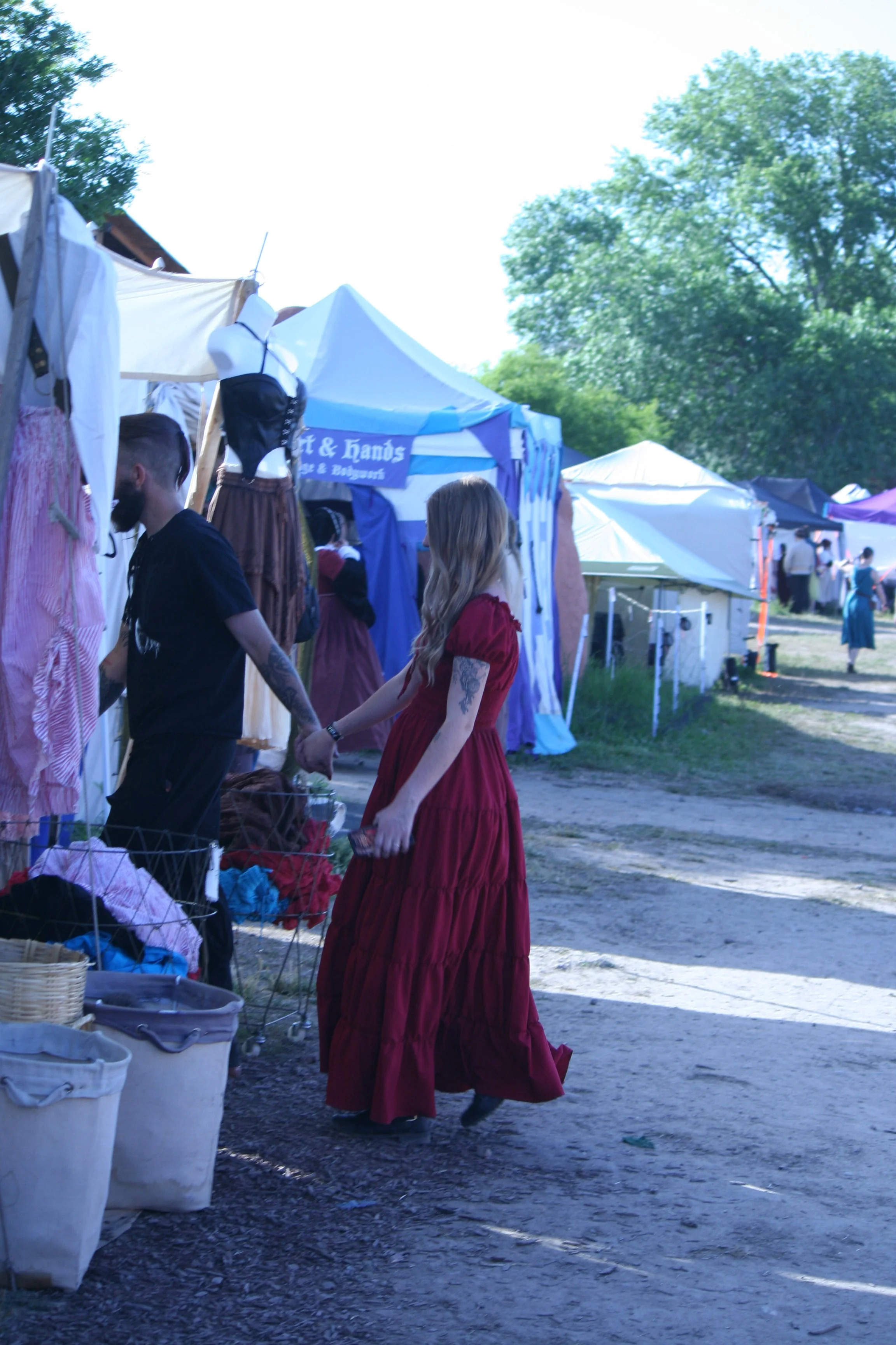 People dressed in vintage clothing holding hands at an outdoor market with tents and trees.