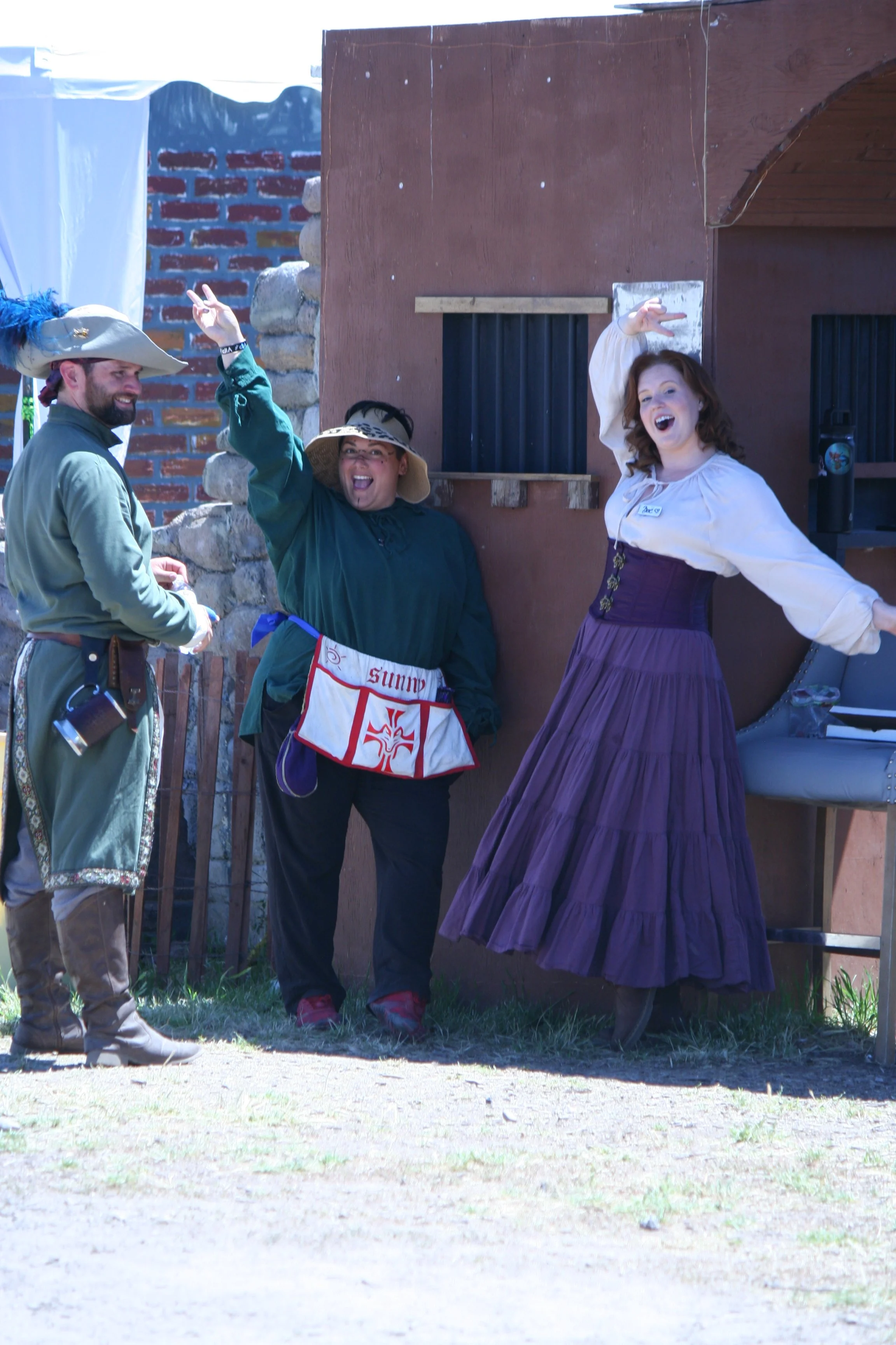 Three people dressed in historical costumes—one man in a pirate costume, one woman in a vintage dress, and another woman in a girl in a green dress with a large sun hat—posing and smiling outdoors near a brown wooden building with small barred window