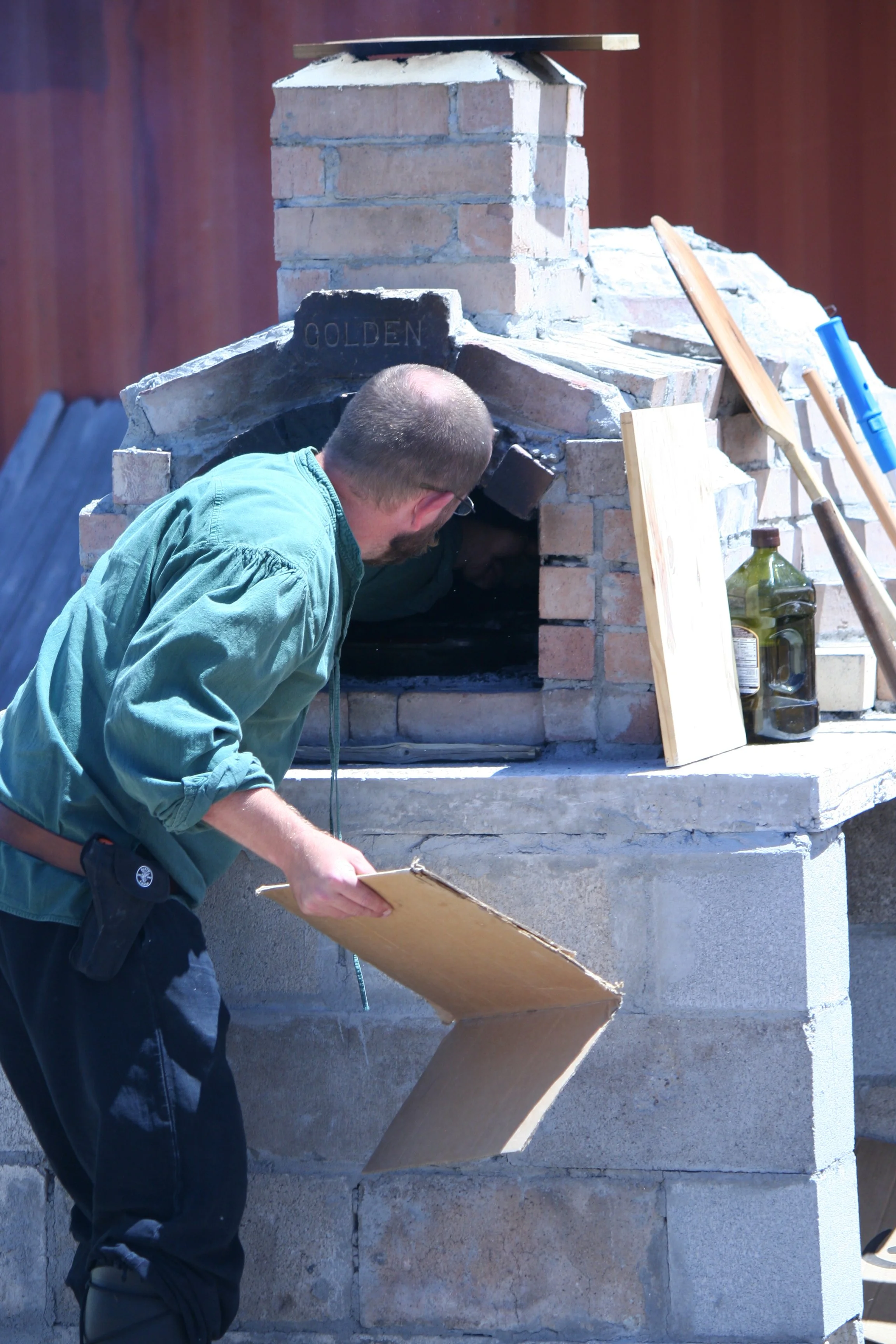 A person with short hair, glasses, and a beard wearing a green jacket and black pants is working on a brick outdoor fireplace. The fireplace has a chimney, and baking tools like paddles are leaning against it. The person is holding a clipboard and is