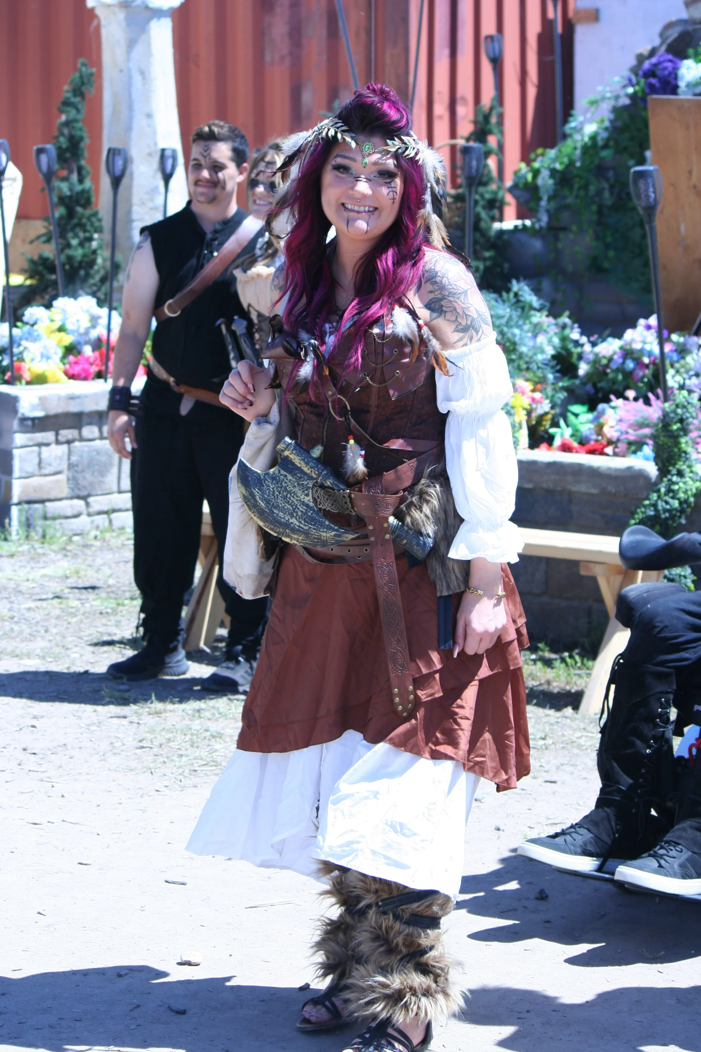 A woman dressed as a Viking with purple hair, face paint, a leafy headband, and furry accessories, standing outdoors in front of a group of people in costume, with colorful flowers and wooden fencing in the background.