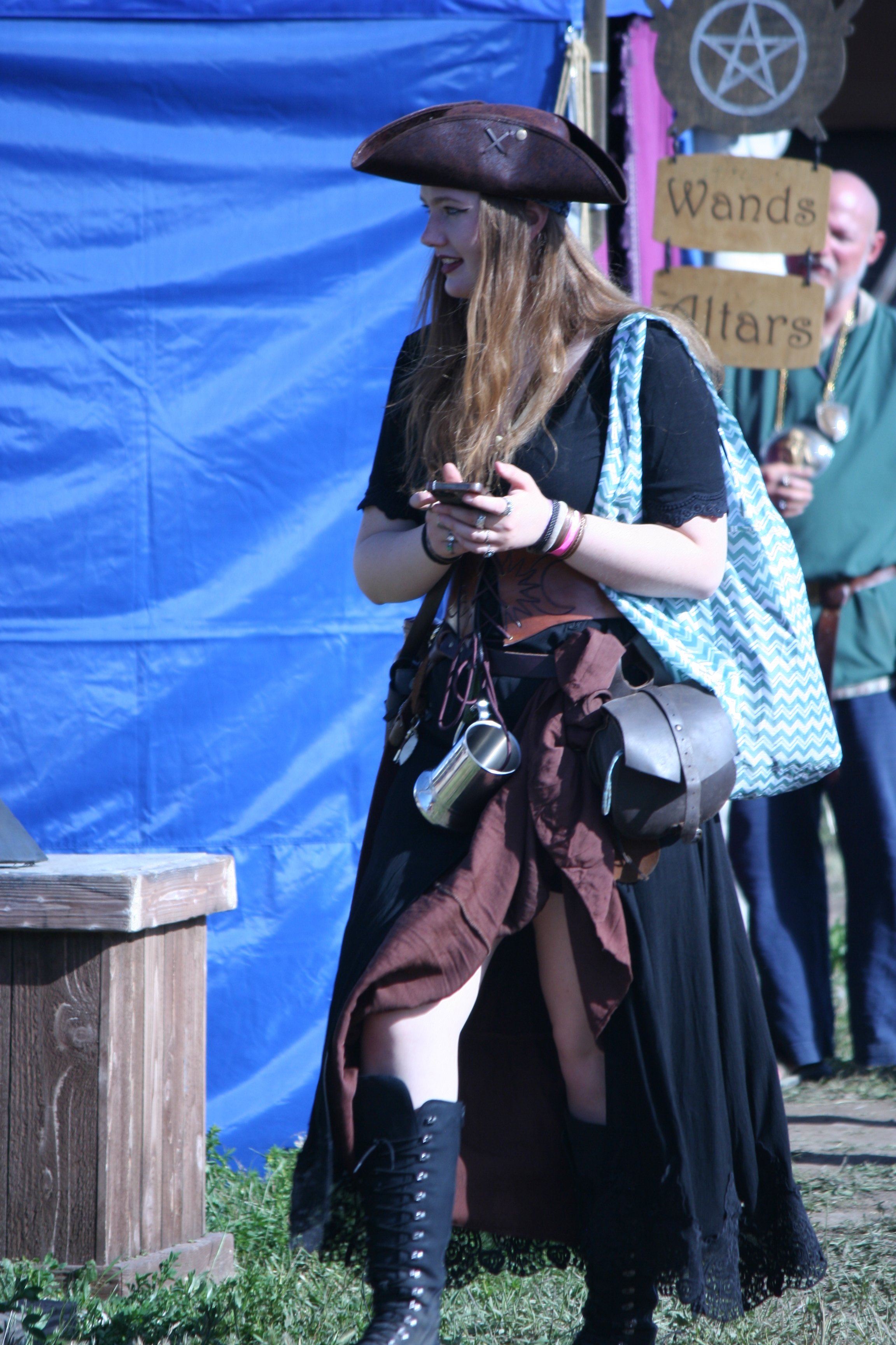 A woman dressed as a pirate in black with a brown cape and lace-up black boots, holding a smartphone, at a fantasy or cosplay event with a blue backdrop and a sign that reads 'Wands' and 'Altar' in the background.