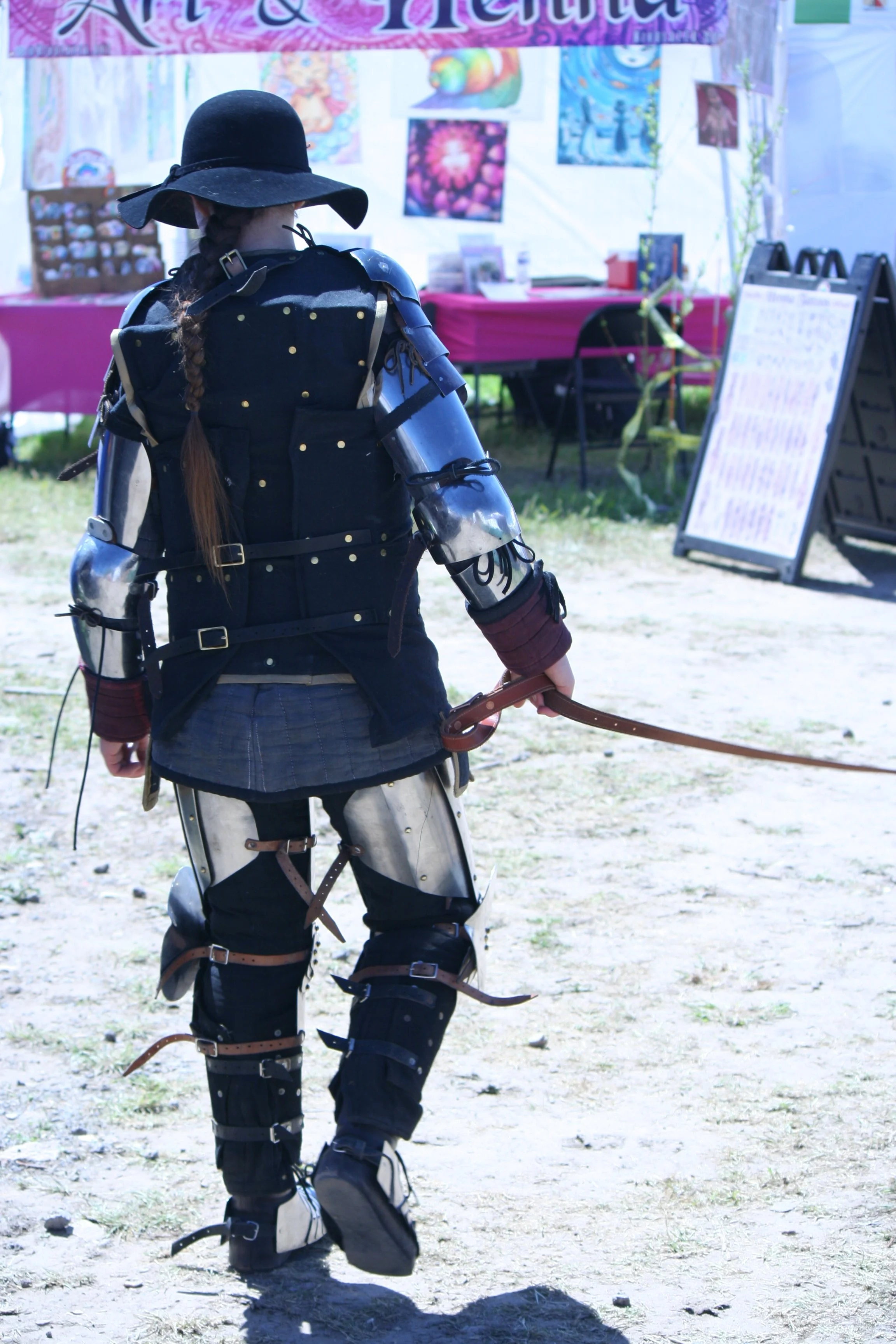A person dressed in a medieval knight costume, including armor, a wide-brimmed black hat, and a long braid, walking outdoors near a booth with colorful posters and a banner.