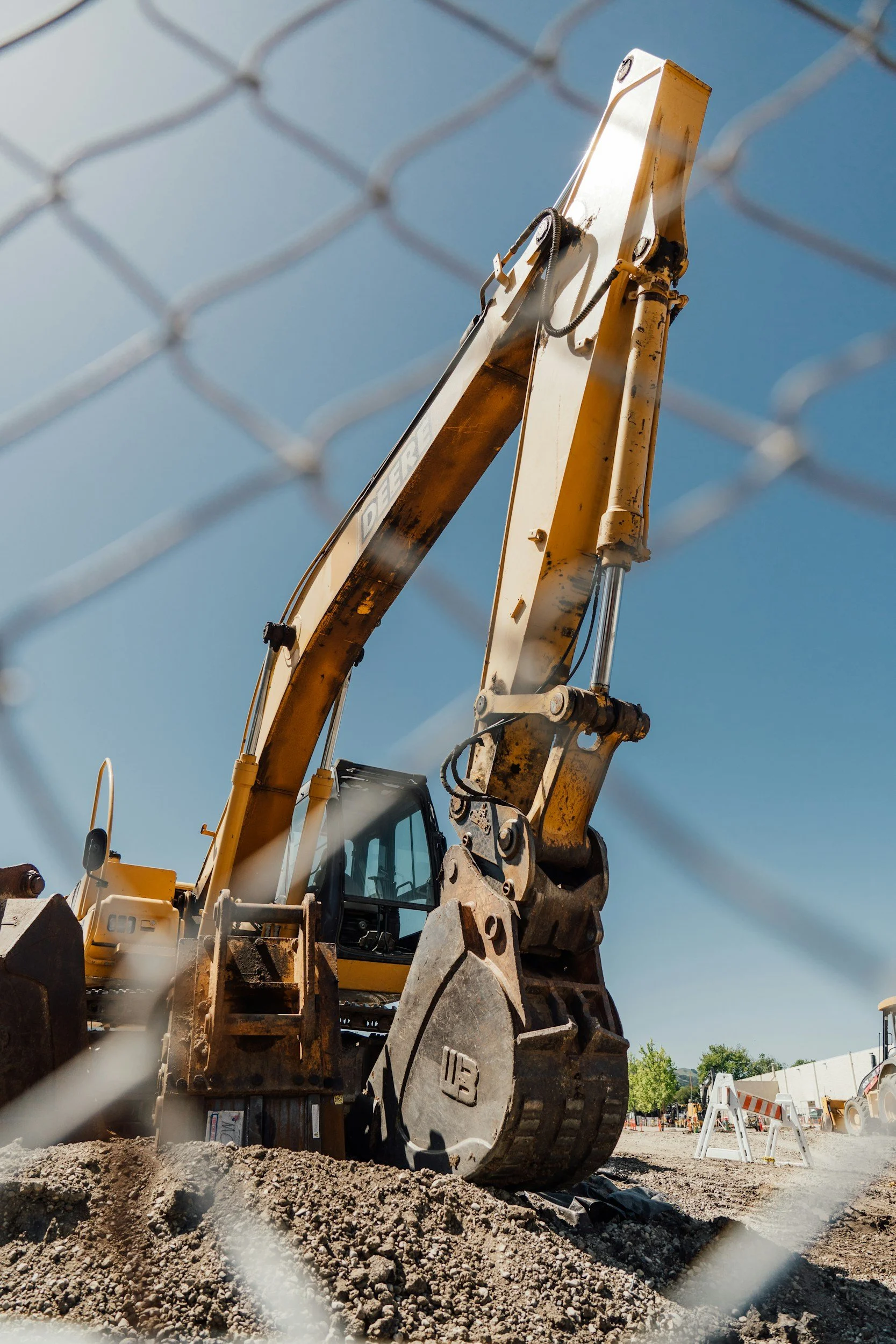 Construction excavator seen through a chain-link fence at a construction site, with a clear blue sky in the background.